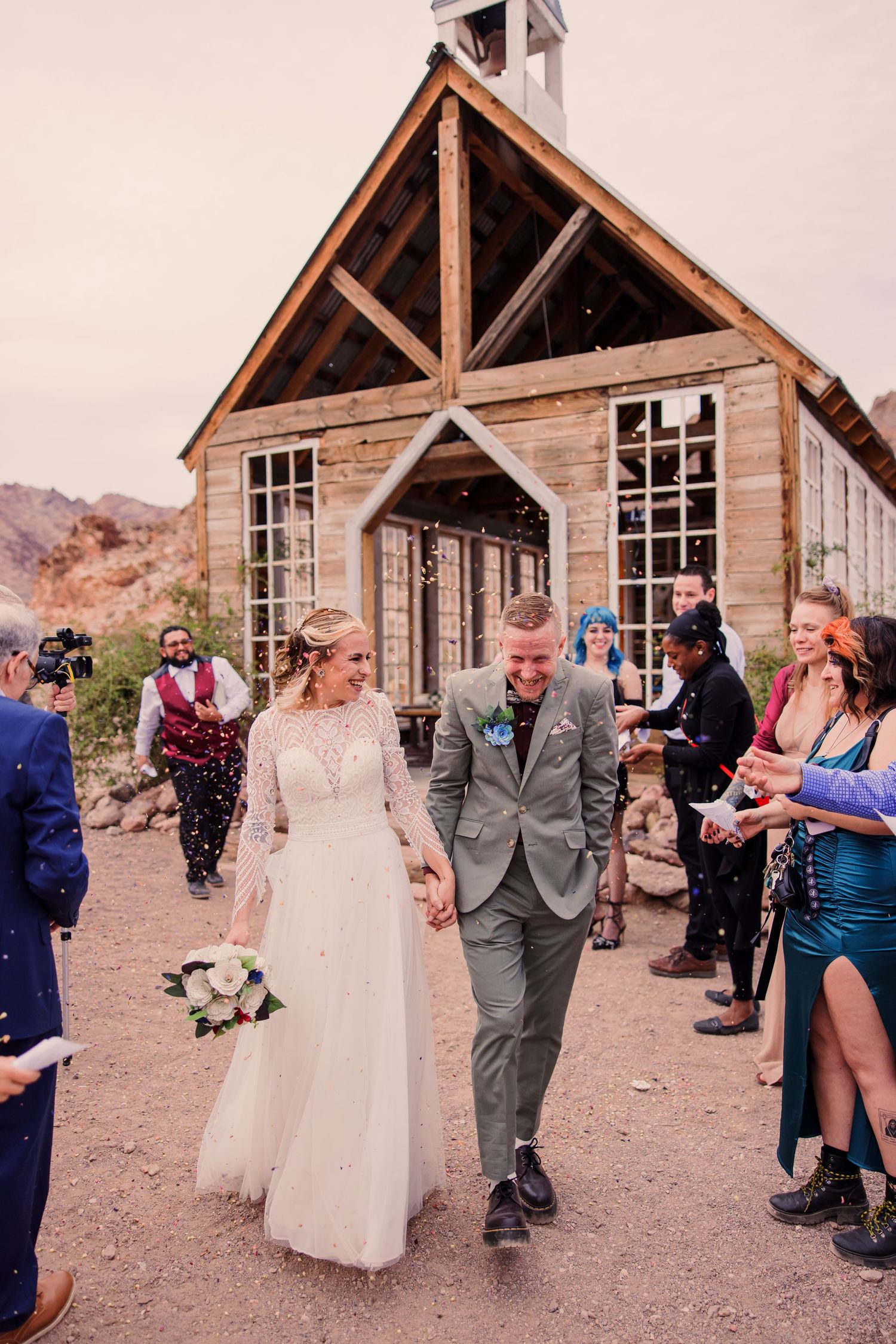 Wedding party celebrates with confetti toss outside rustic wooden church building in desert landscape setting.