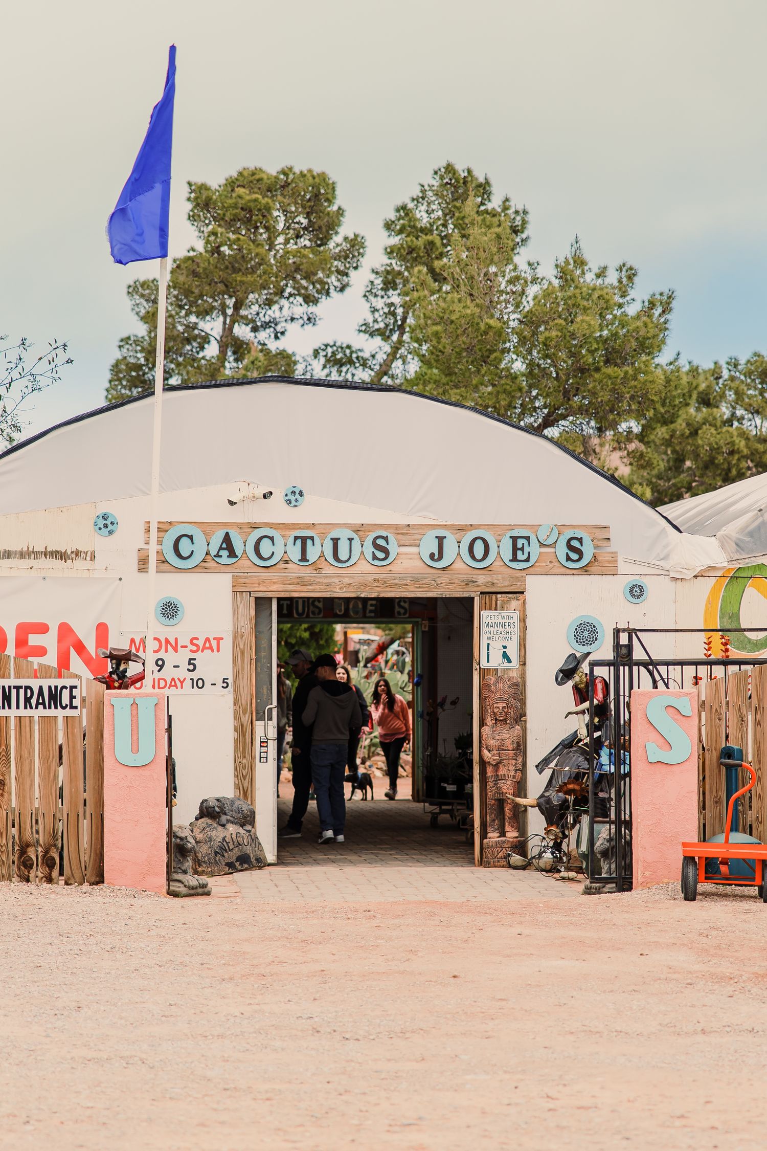 Panoramic view of a rustic desert storefront with pink walls and blue signs reading 'Cactus Joe's' against a cloudy sky.