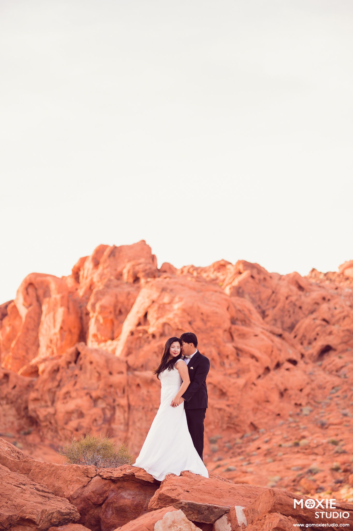 A wedding couple poses on red rock formations in a desert landscape during golden hour.
