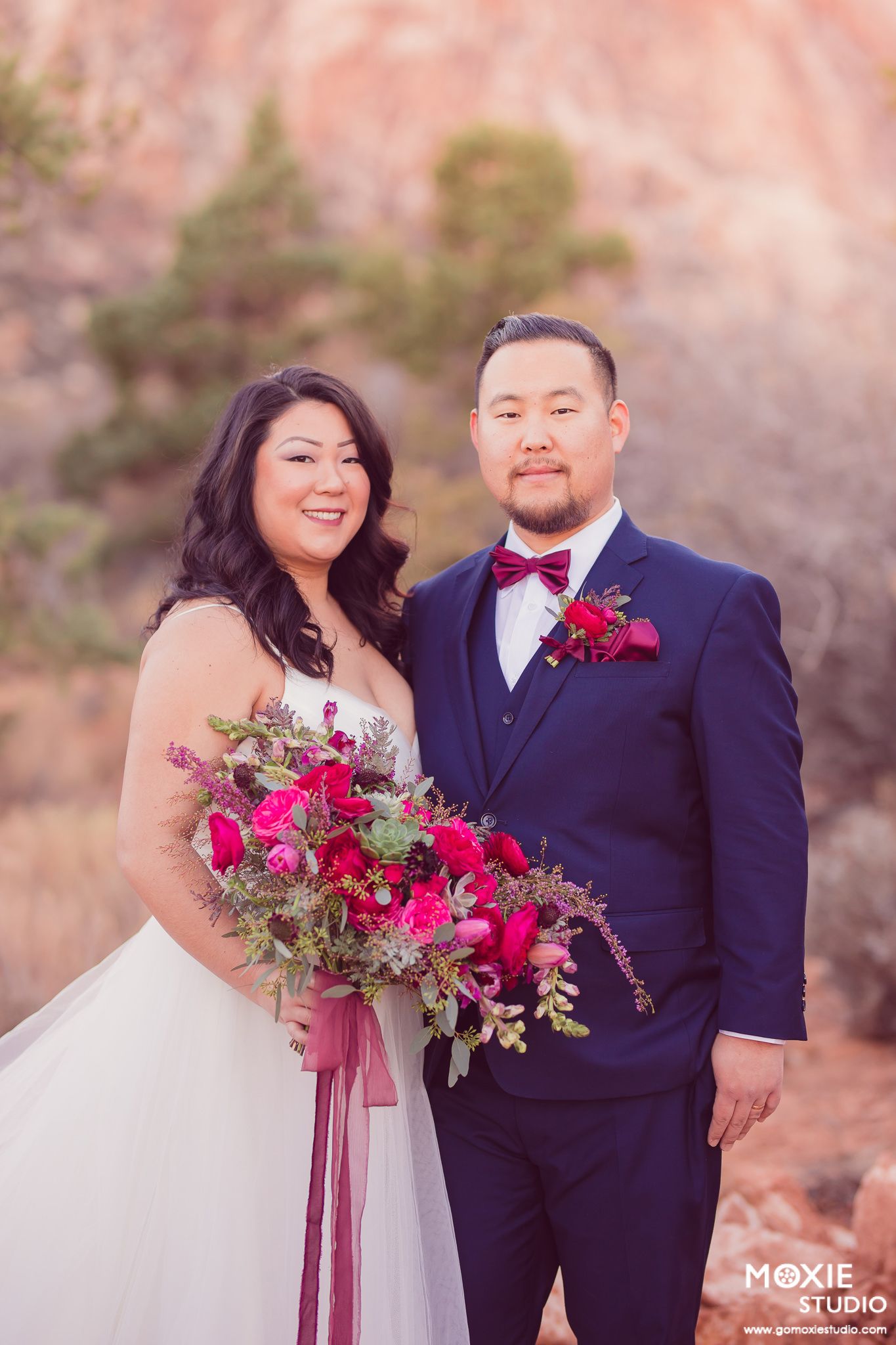 A romantic outdoor wedding photoshoot featuring a couple in navy and white attire with vibrant red floral bouquets.