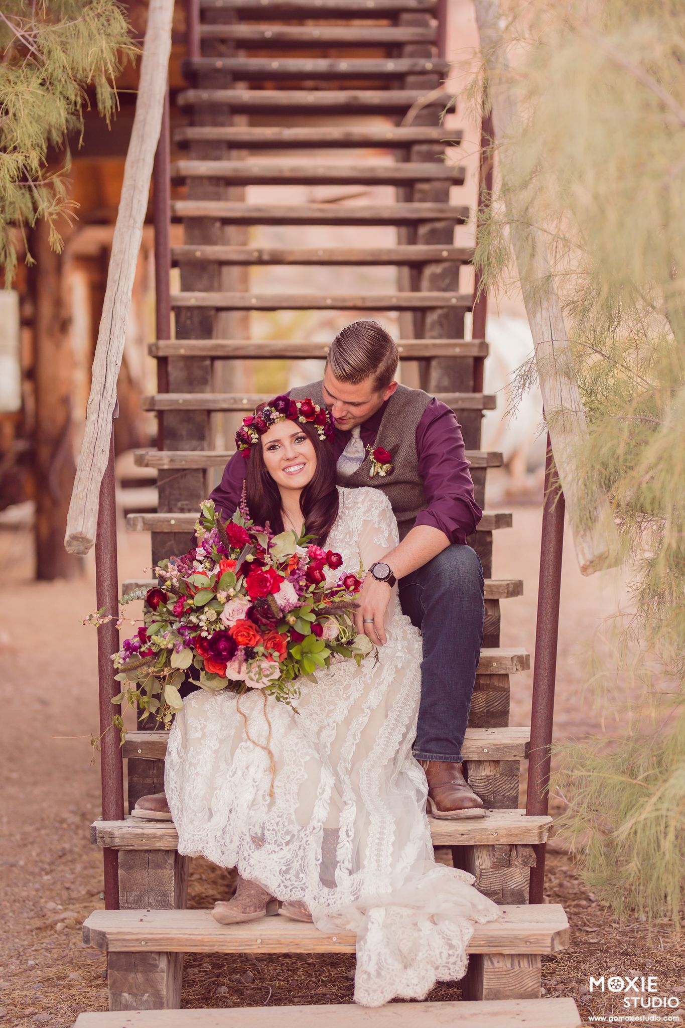 A couple sits together on rustic wooden stairs surrounded by romantic floral arrangements in deep burgundy and red tones.