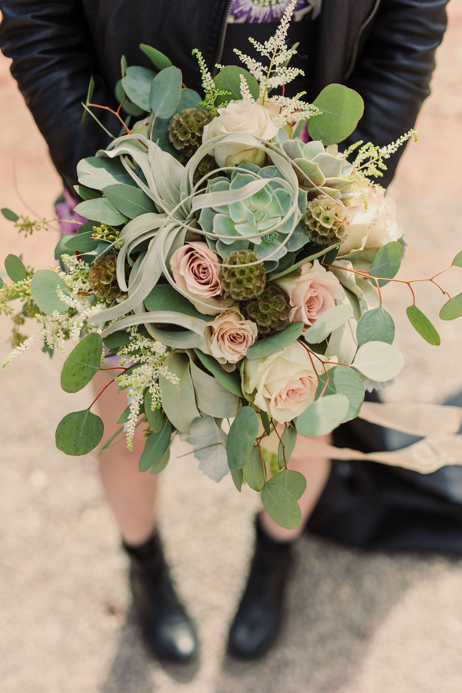 Elegant wedding bouquet with blush roses, eucalyptus, and succulents held against a black suit.