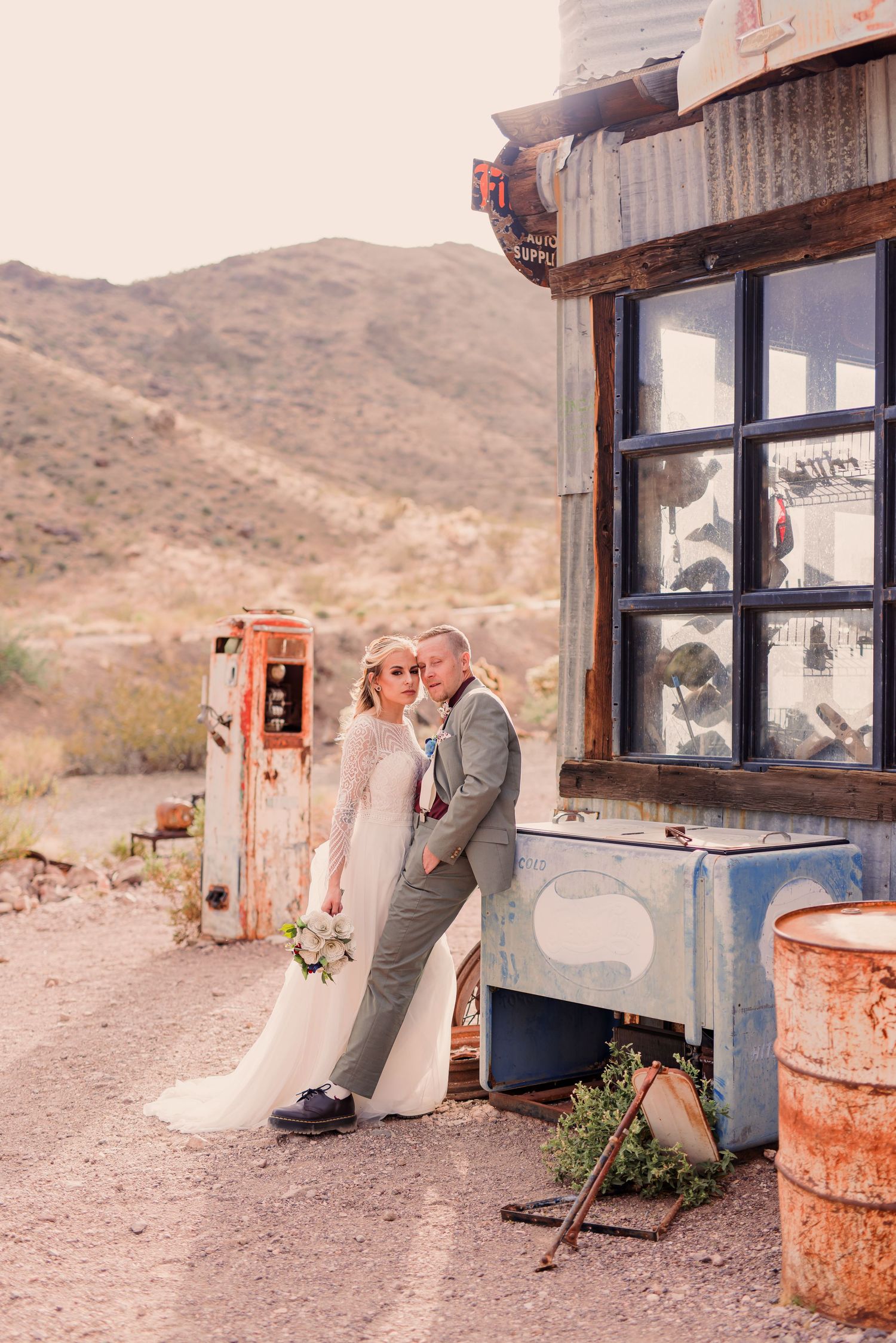 A couple in wedding attire poses together at a rustic desert gas station with vintage fuel pumps and mountains in background.