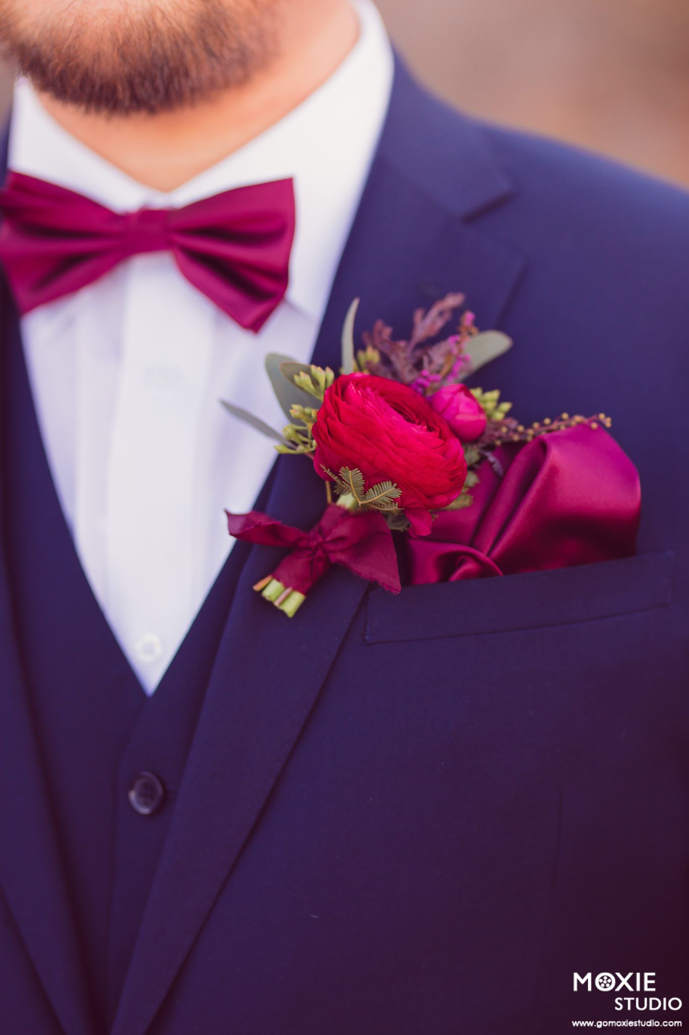 A burgundy bow tie and red rose boutonniere on a navy blue suit jacket lapel.