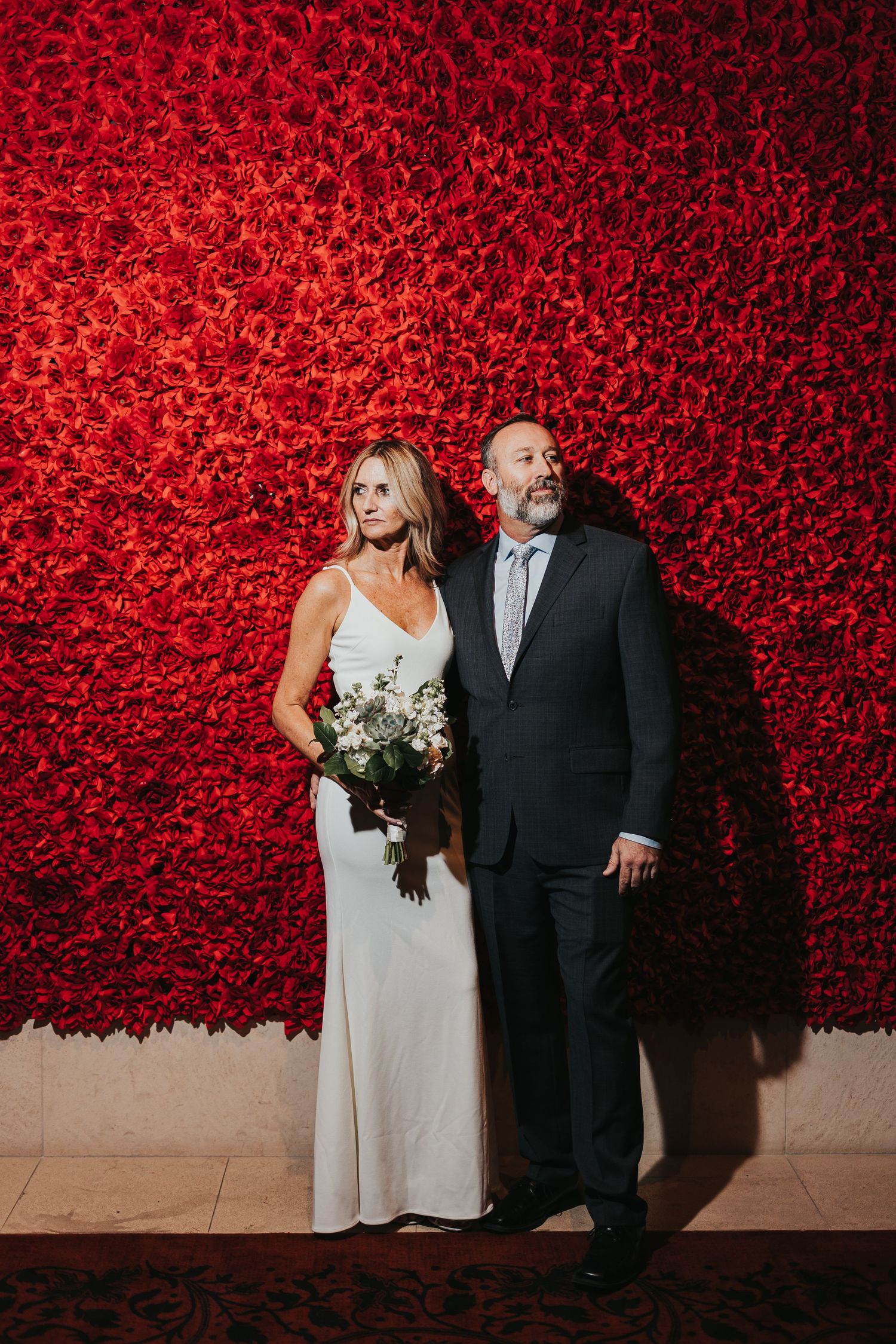 Wedding couple poses against stunning backdrop of bright red rose wall in sequence of candid moments.