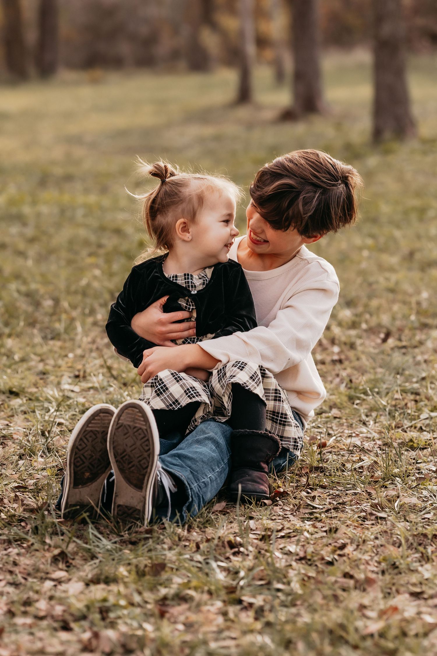 Brother and sister smiling at each other while sitting among colorful fall leaves during a family photo session