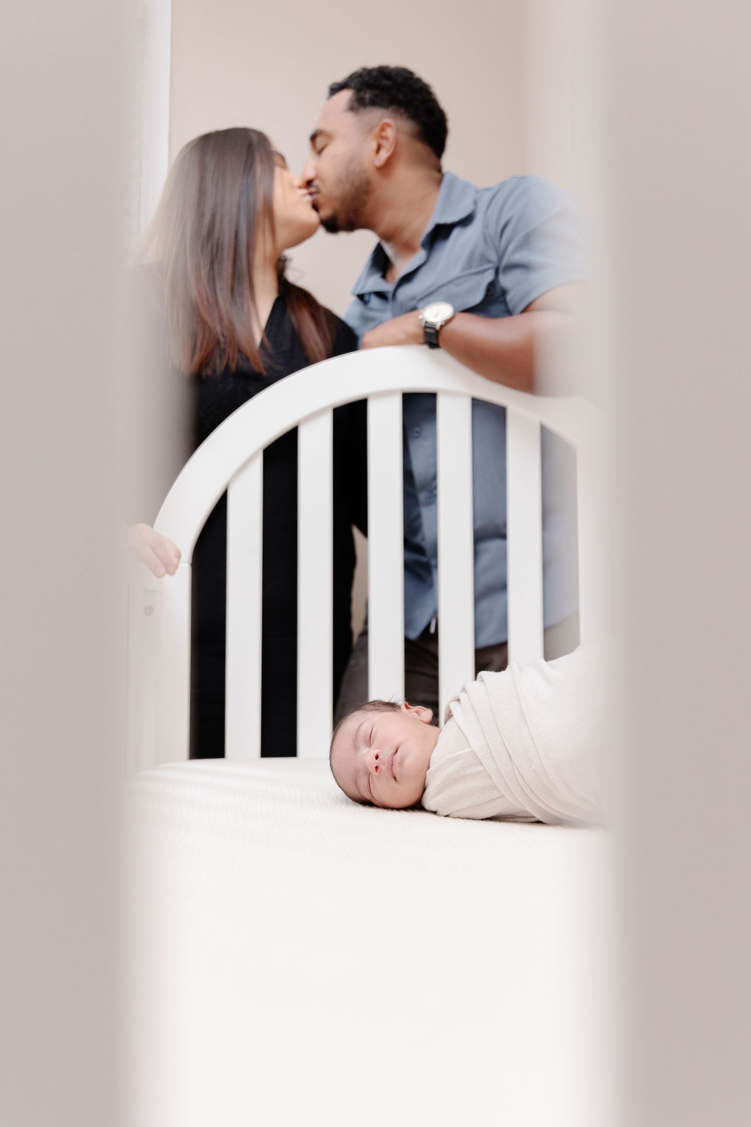 Newborn baby swaddled in crib while parents share a kiss in the background during a family photo session.