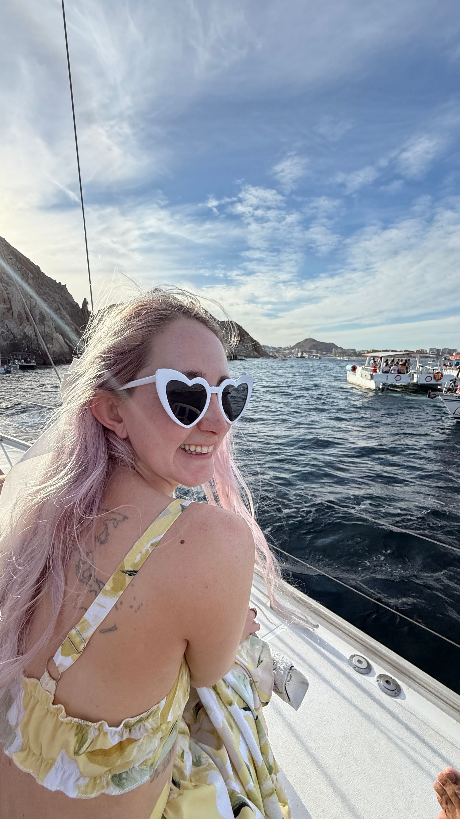 Woman with pink hair and white heart sunglasses smiling on a sailboat with rocky coastline and blue ocean behind her.