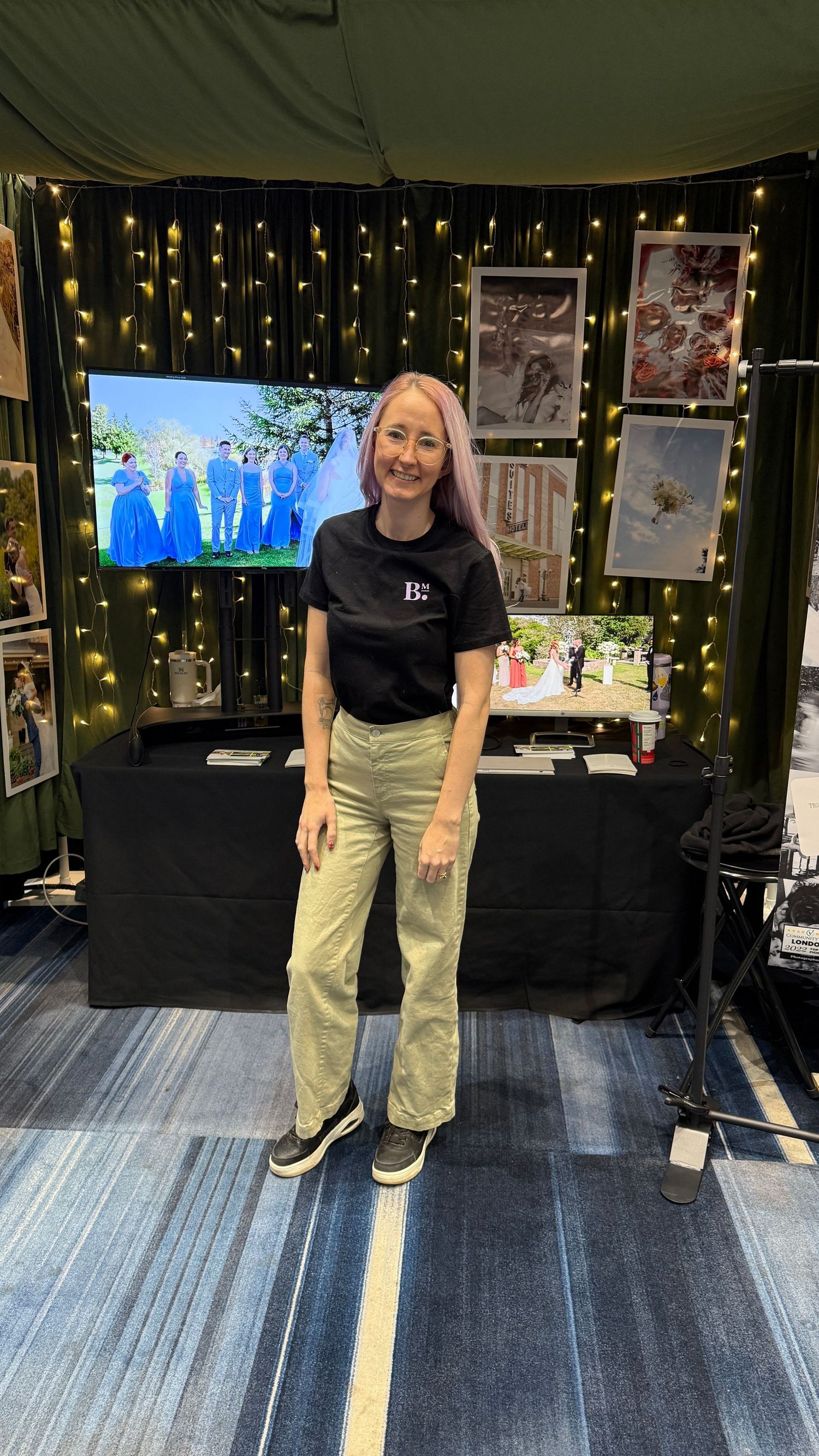Woman with pink hair in black tee and khaki pants stands before a photography booth with string lights and wedding photos.