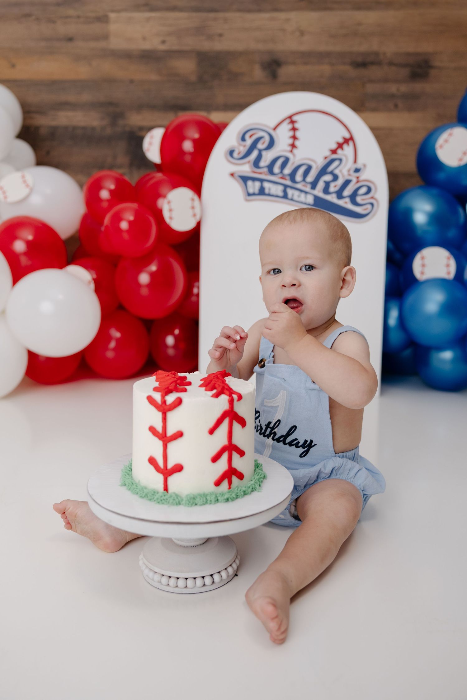 Baby boy in blue romper touching baseball-themed first birthday cake surrounded by red, white, and blue balloons.