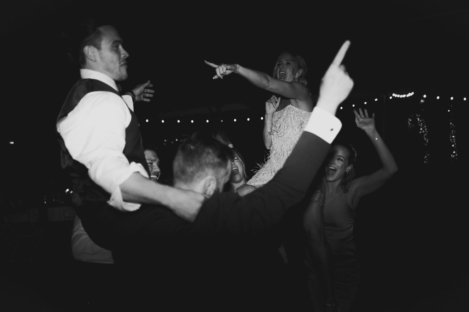 Black and white photo of wedding guests dancing joyfully at a lively outdoor evening reception.