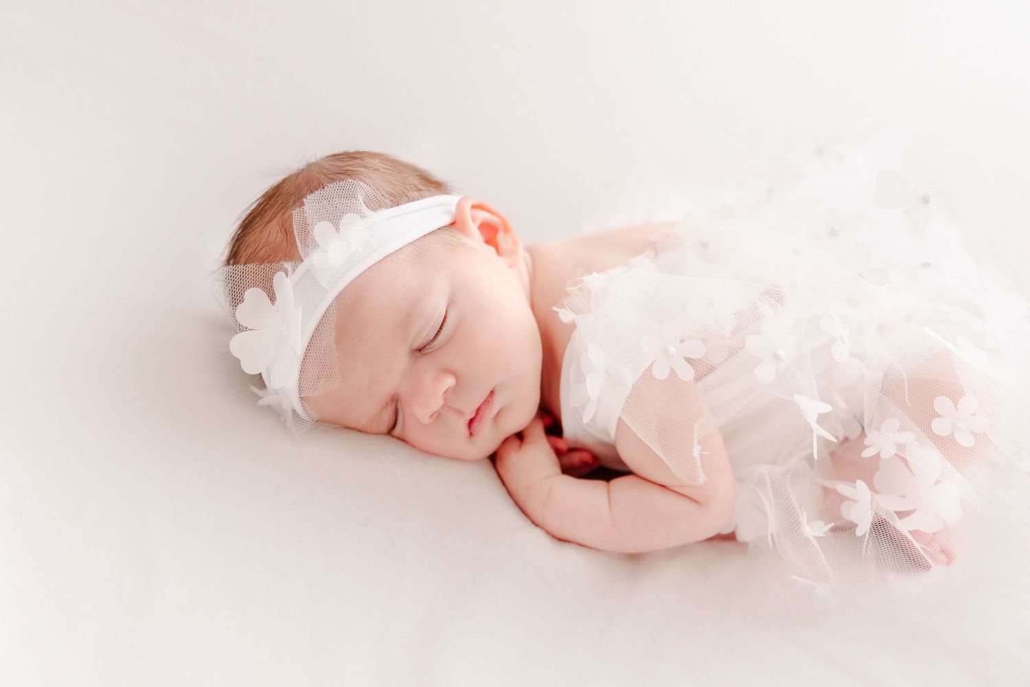 Sleeping newborn baby girl in white tulle dress and floral headband on soft white background.