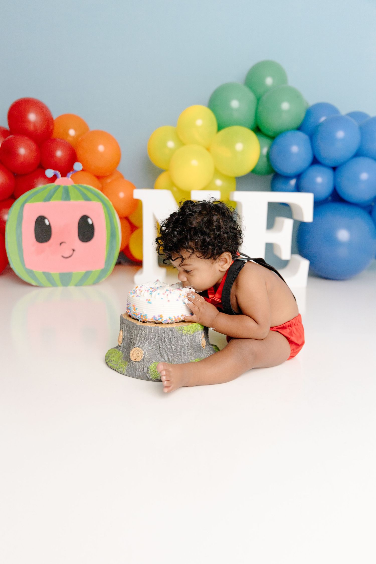 Cake smash photo with rainbow balloon arch and watermelon decoration for first birthday celebration.