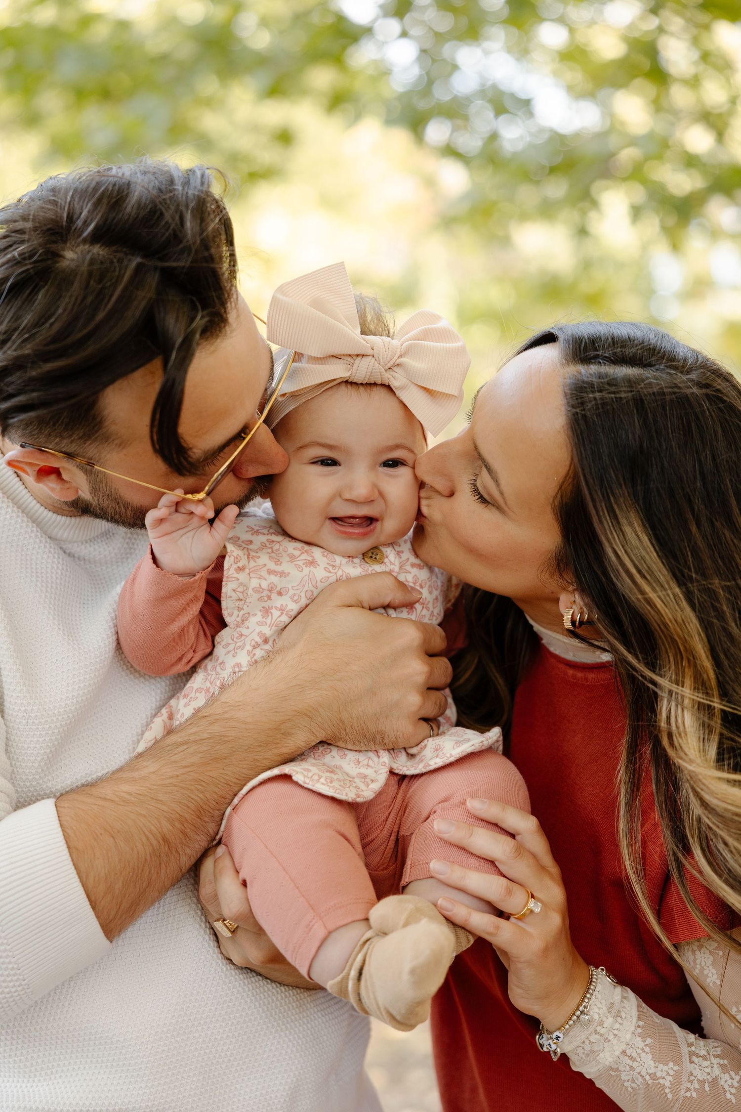 A family shares tender moments and kisses with their baby in a series of outdoor portraits with soft natural lighting.