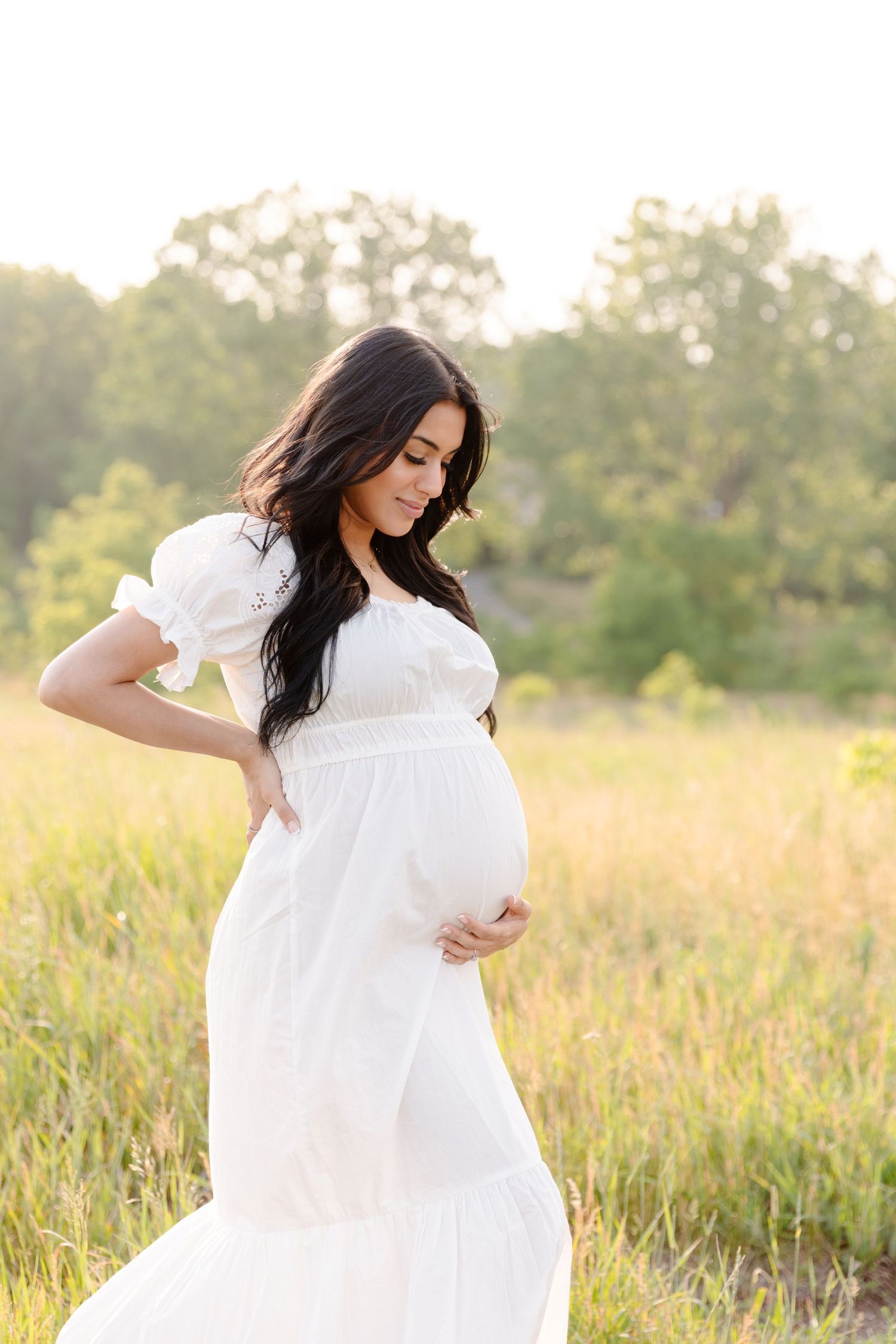 A woman in a flowing white dress stands in a golden sunlit field during a maternity photoshoot.