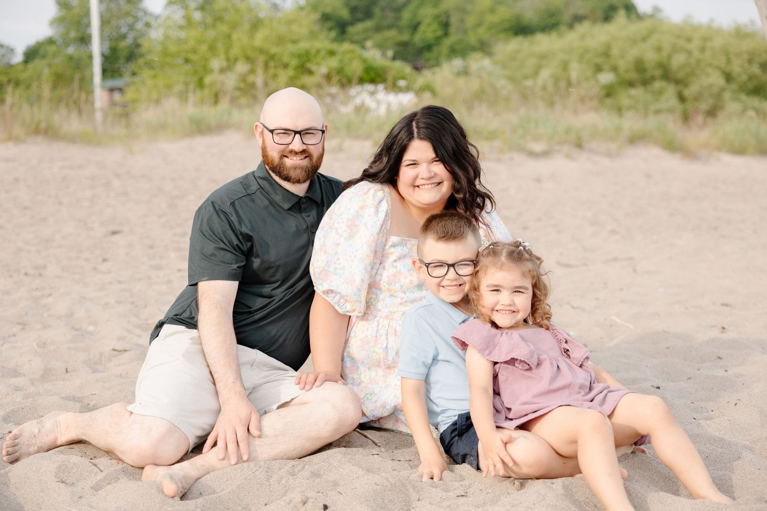 A family sits together on a sandy beach wearing casual clothing on a sunny day.