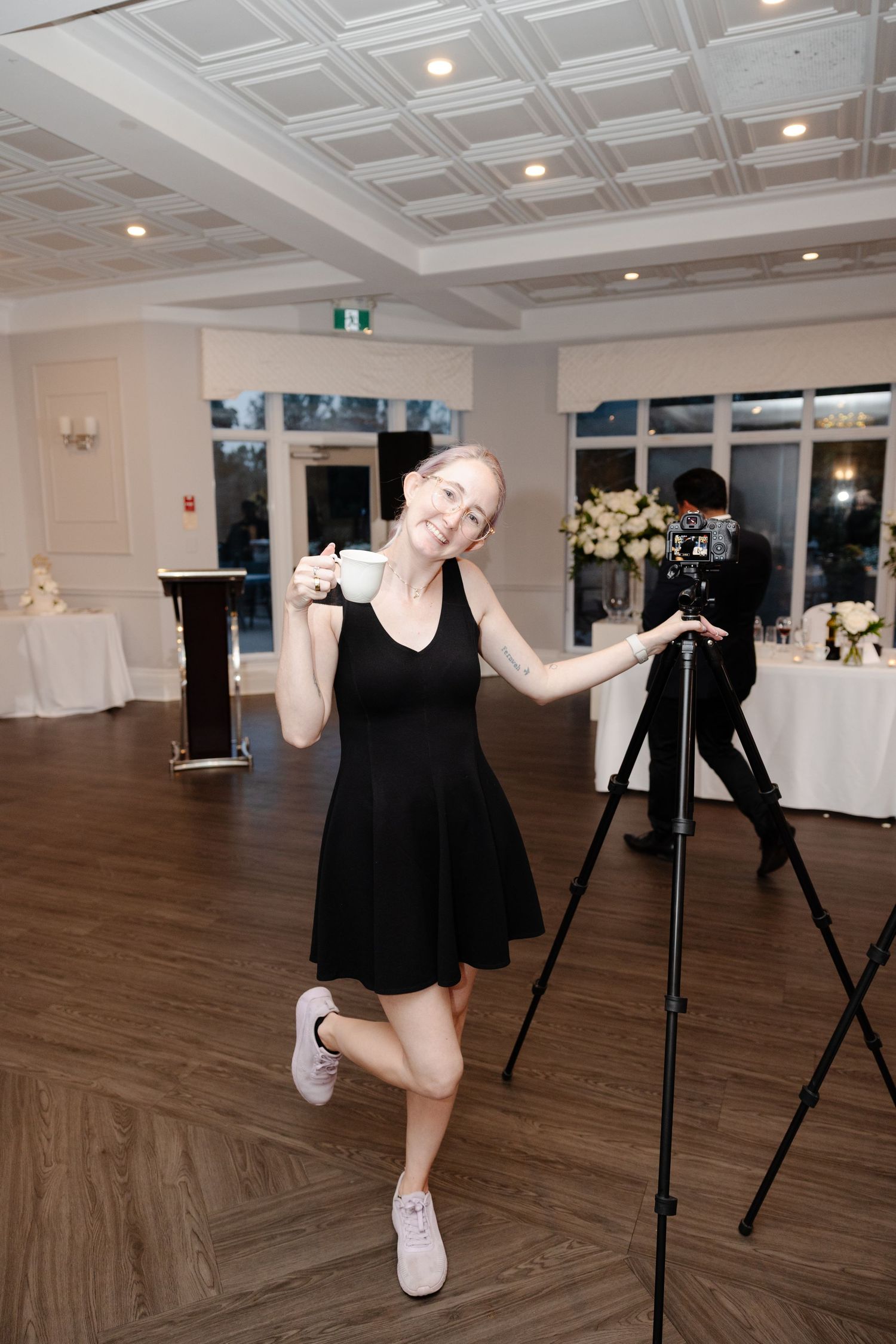 Woman in black dress poses playfully with coffee cup near camera tripod in elegant white event venue.