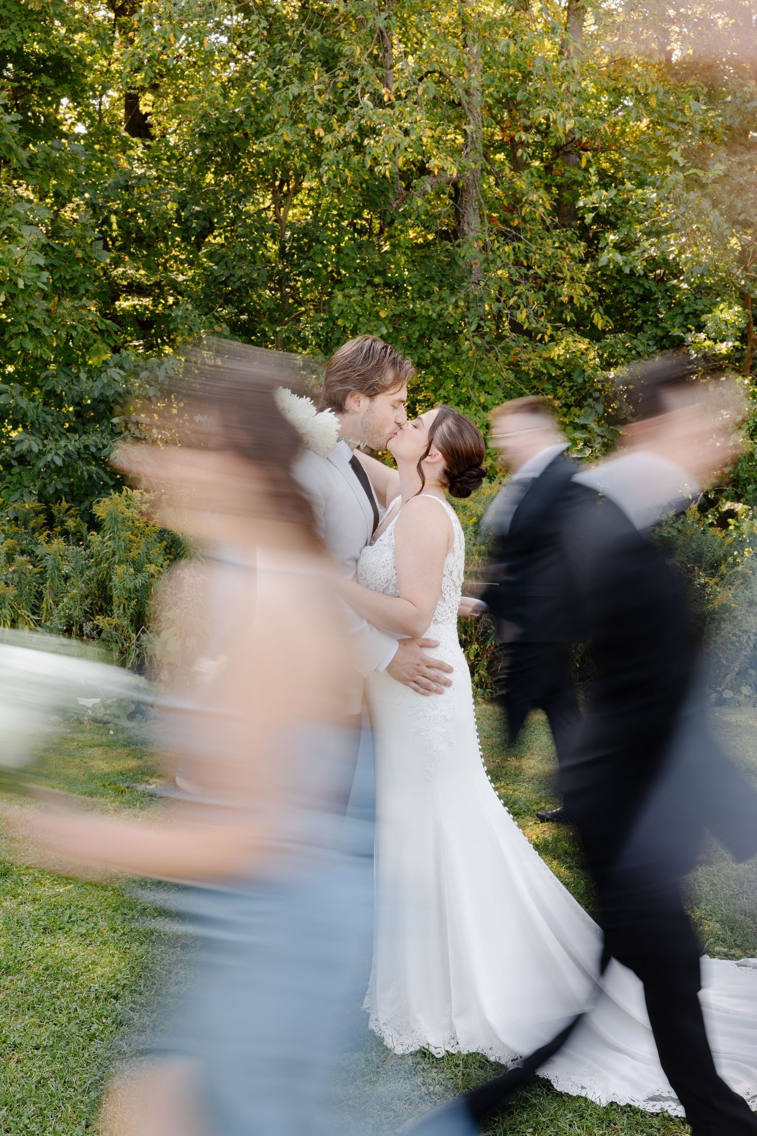Bride and groom share a kiss while wedding party moves around them in a blurred motion outdoors.