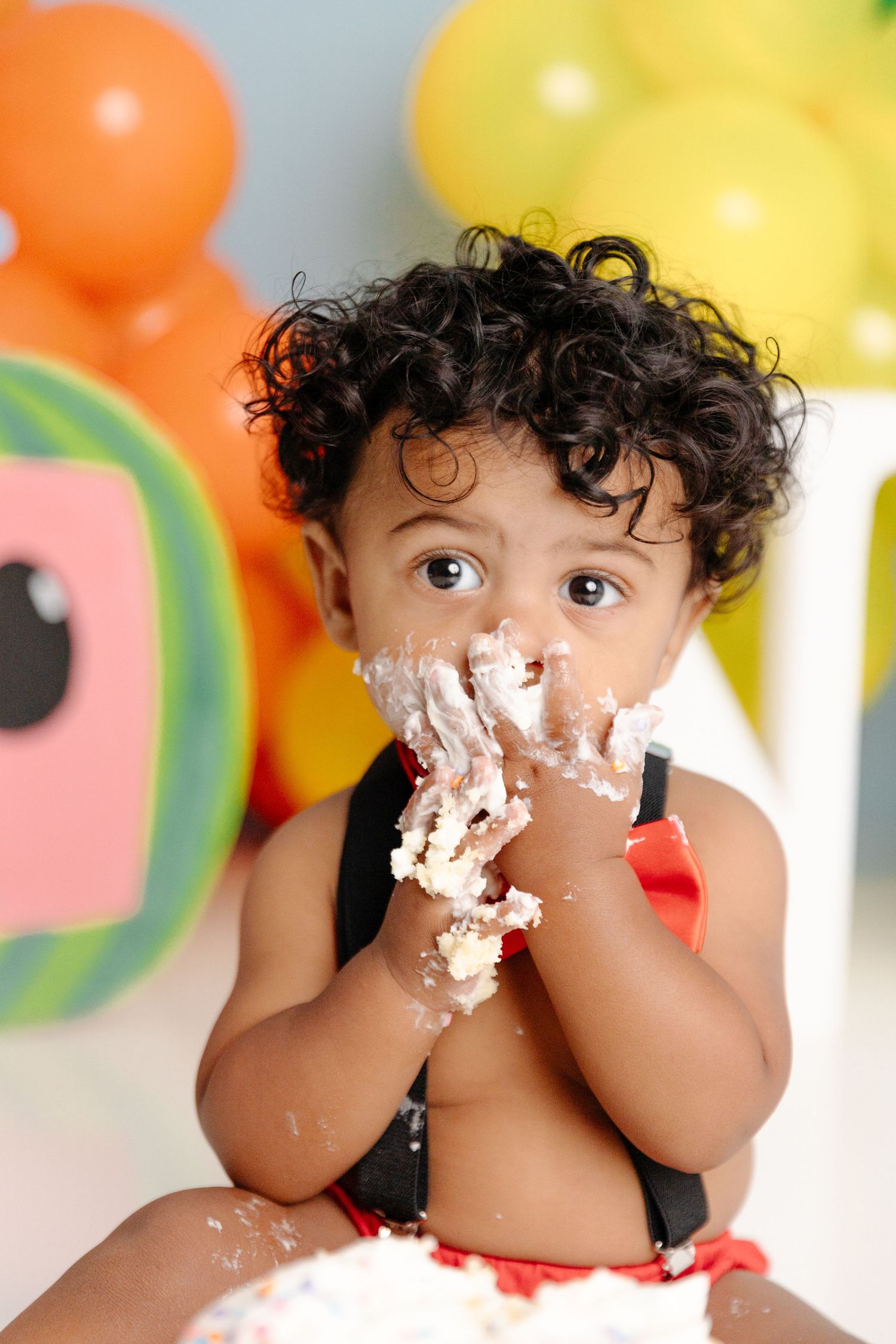 Toddler with curly hair enjoys a cake smash birthday celebration with colorful balloons in the background.