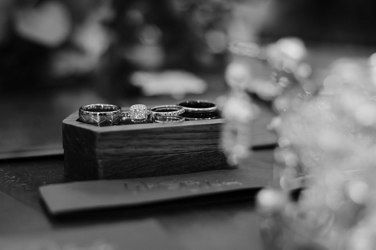 Black and white photo of wedding rings displayed on a small wooden box with floral bokeh background.
