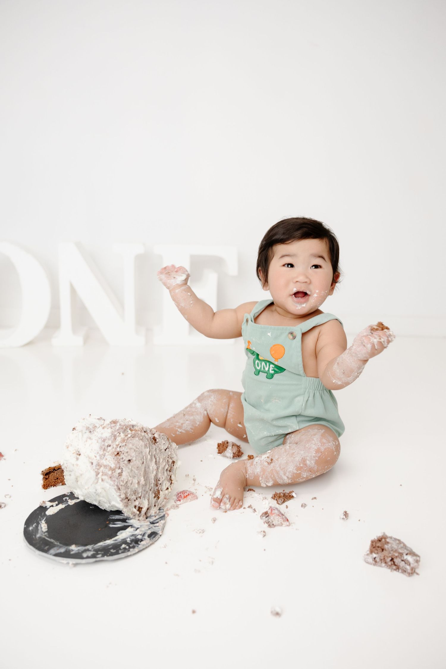Baby in green romper smashes first birthday cake, covered in frosting, sitting in front of ONE sign.