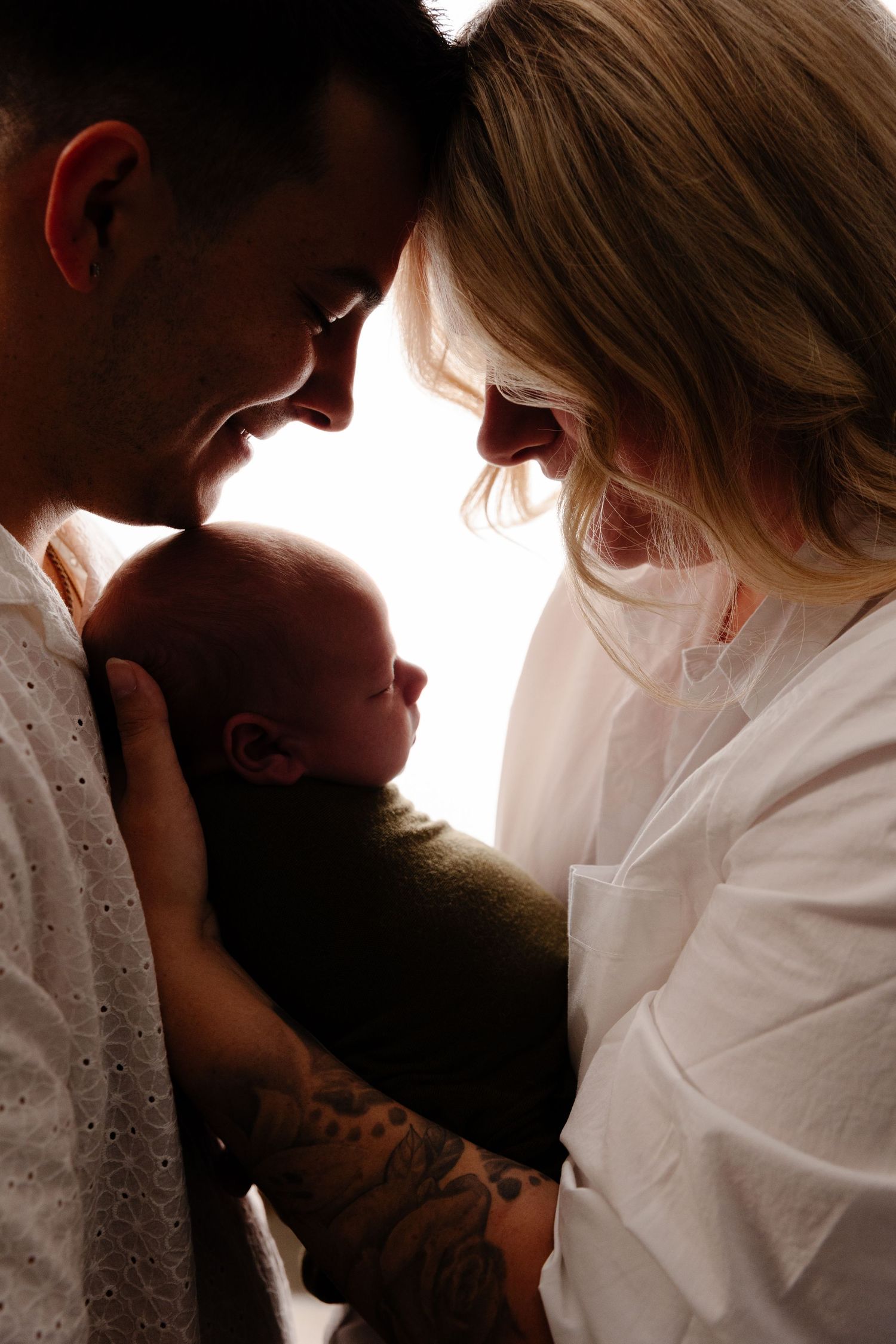 New parents tenderly hold their newborn baby together, father's tattooed arm visible in a backlit portrait.