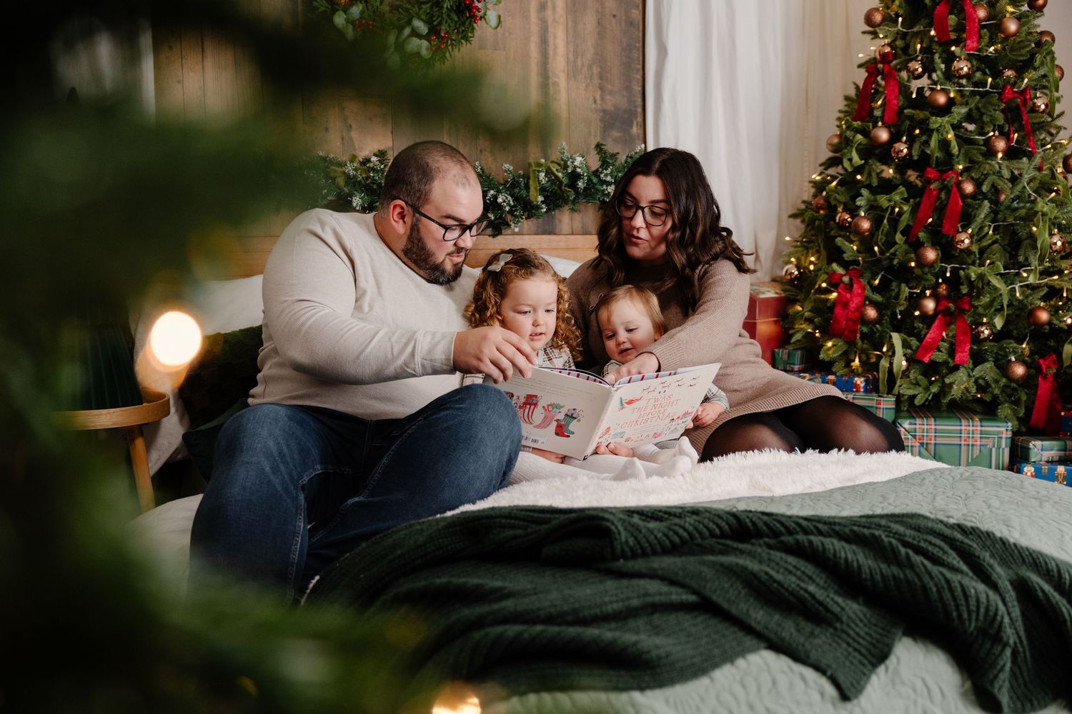 A cozy Christmas scene with people relaxing on a bed near a decorated tree and glowing lights.