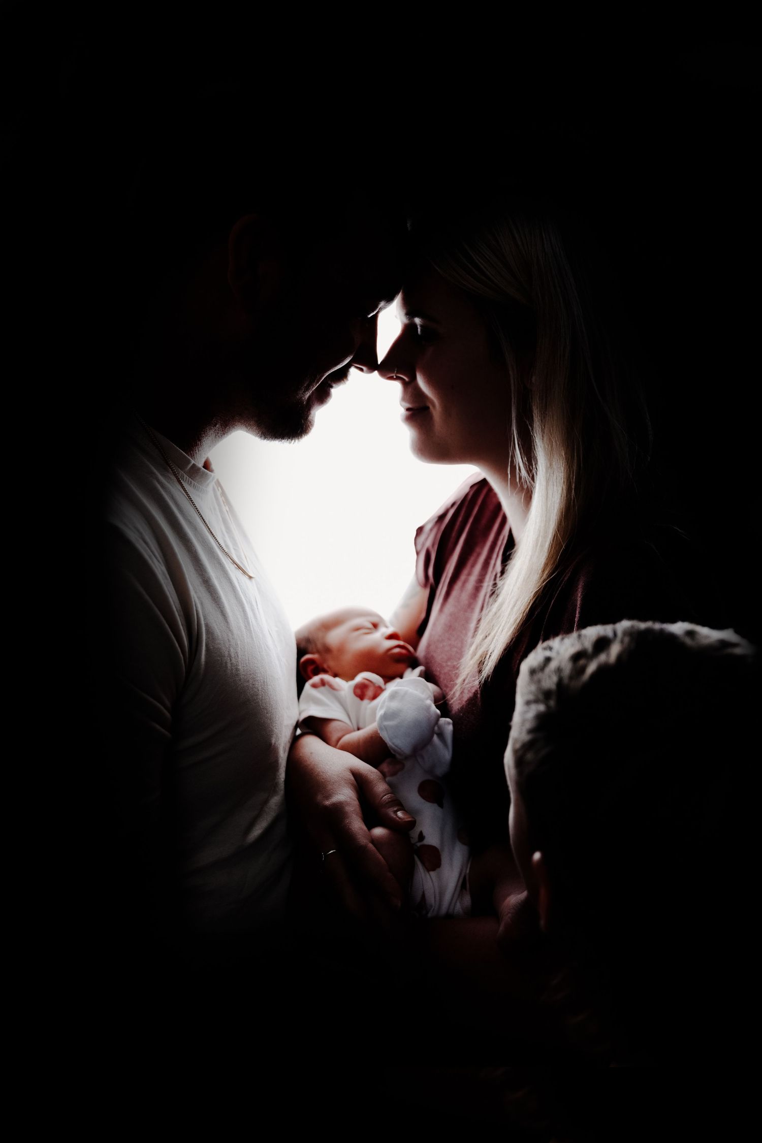 Intimate silhouette portrait of new parents with baby illuminated by soft light against dark background.