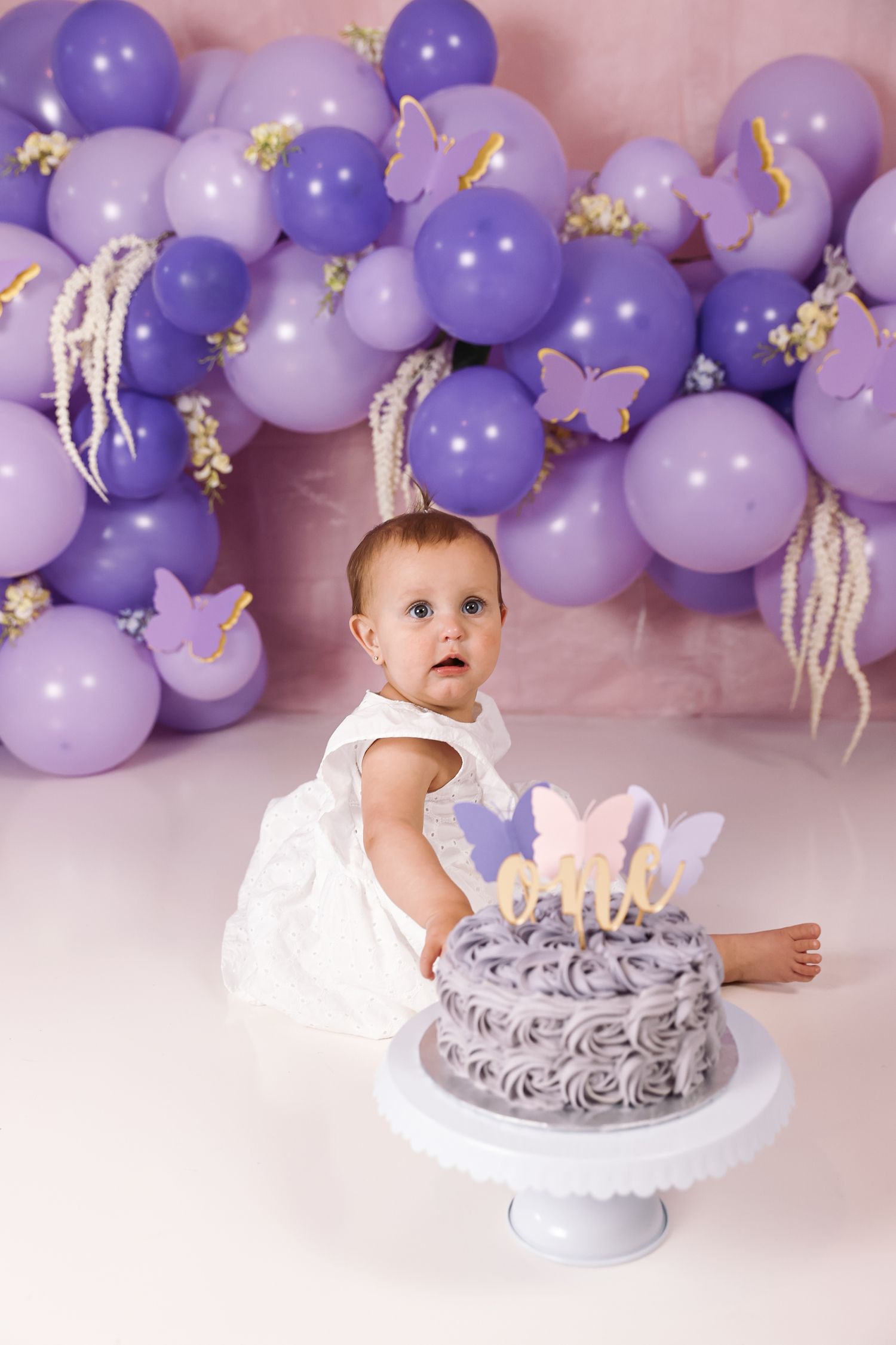 Baby girl in white dress sits before purple rosette smash cake with butterfly topper, surrounded by purple balloon garland.
