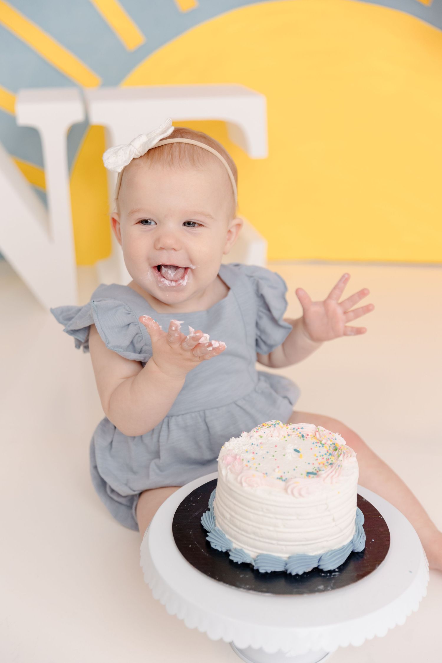 Happy baby girl in gray romper smashing first birthday cake with white frosting and sprinkles on white floor.
