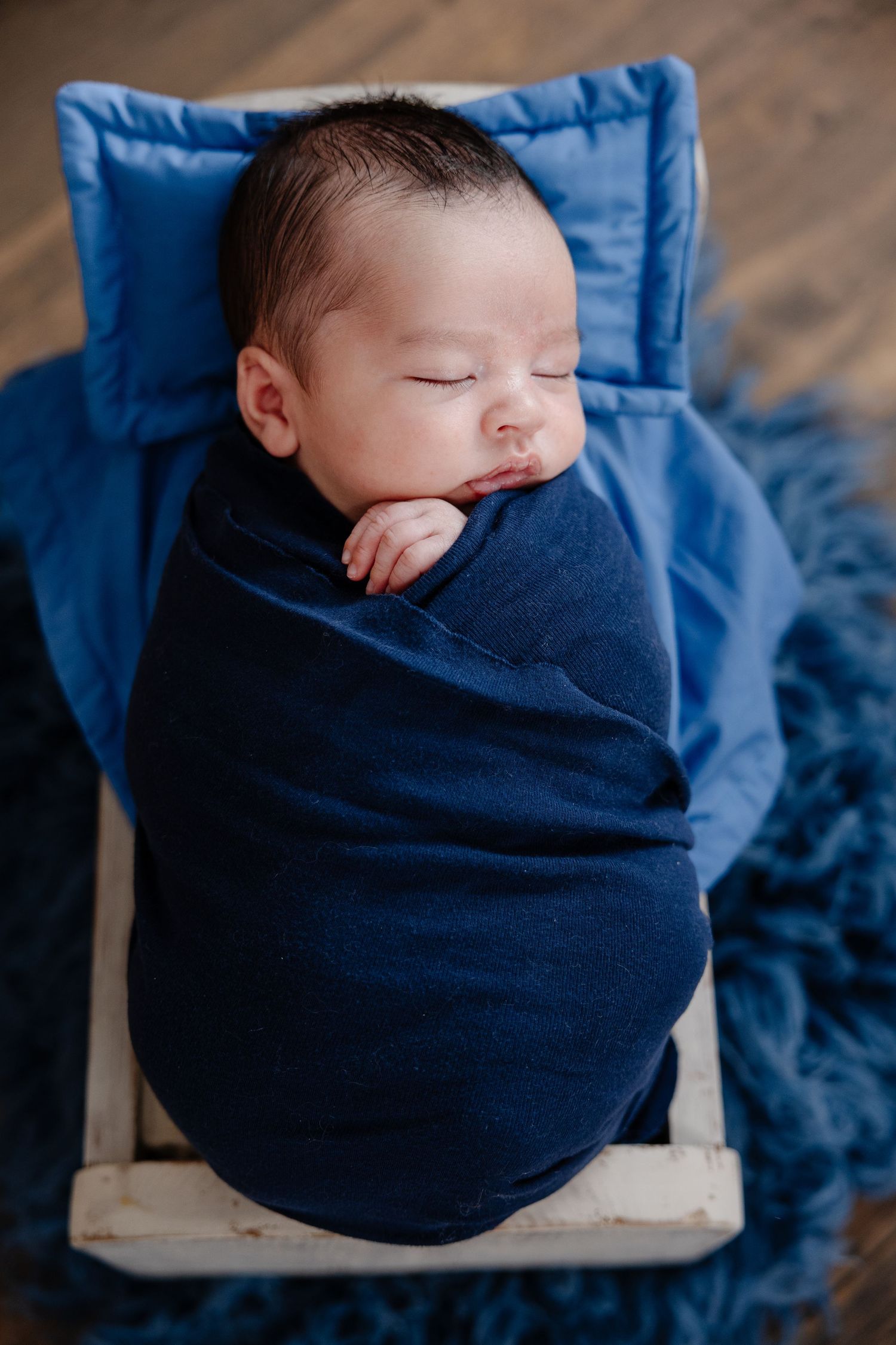 Sleeping newborn baby swaddled in navy blue wrap on miniature wooden bed with blue pillow and fur blanket.