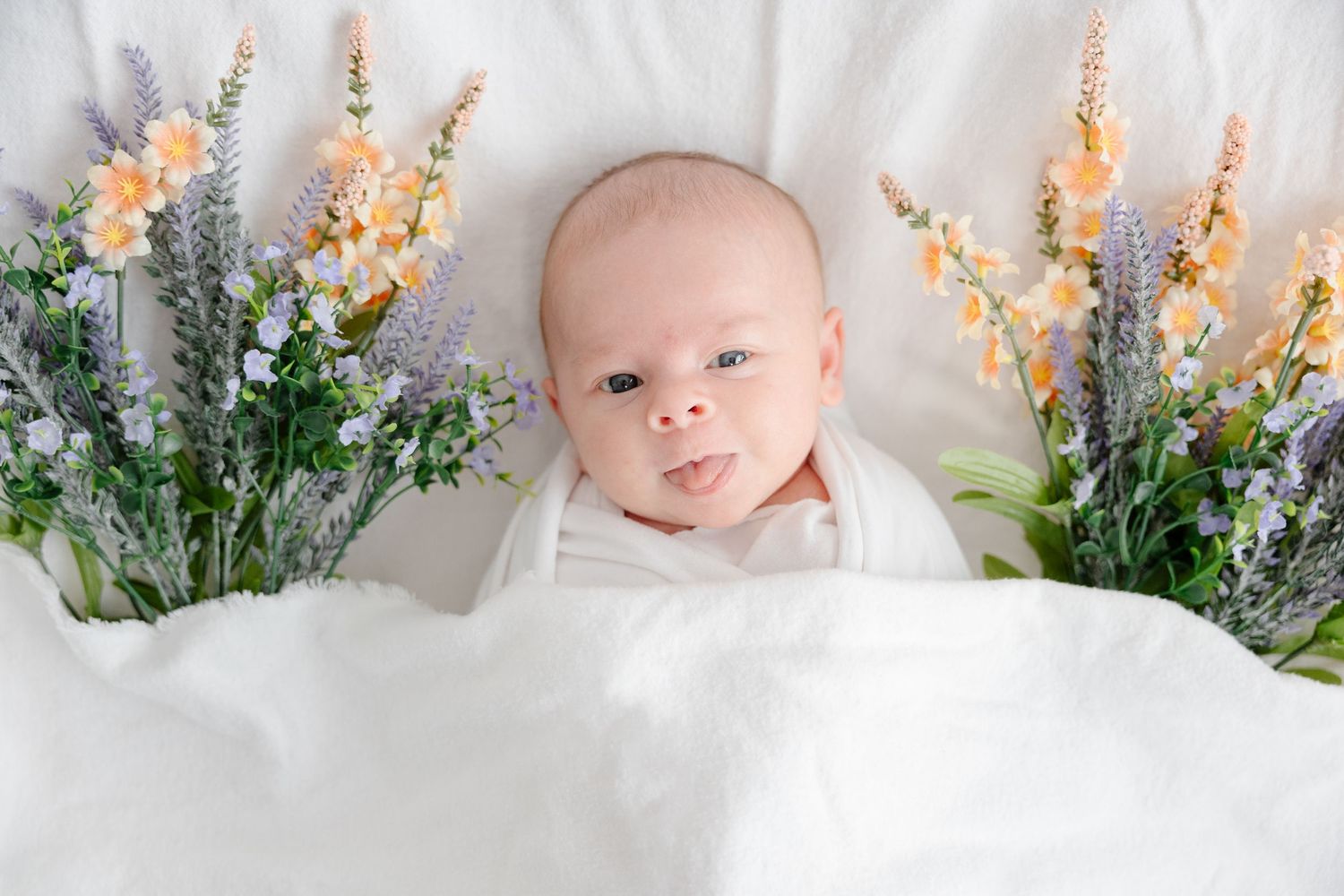 Newborn baby wrapped in white swaddle lying between purple lavender and orange flower arrangements on white blanket.