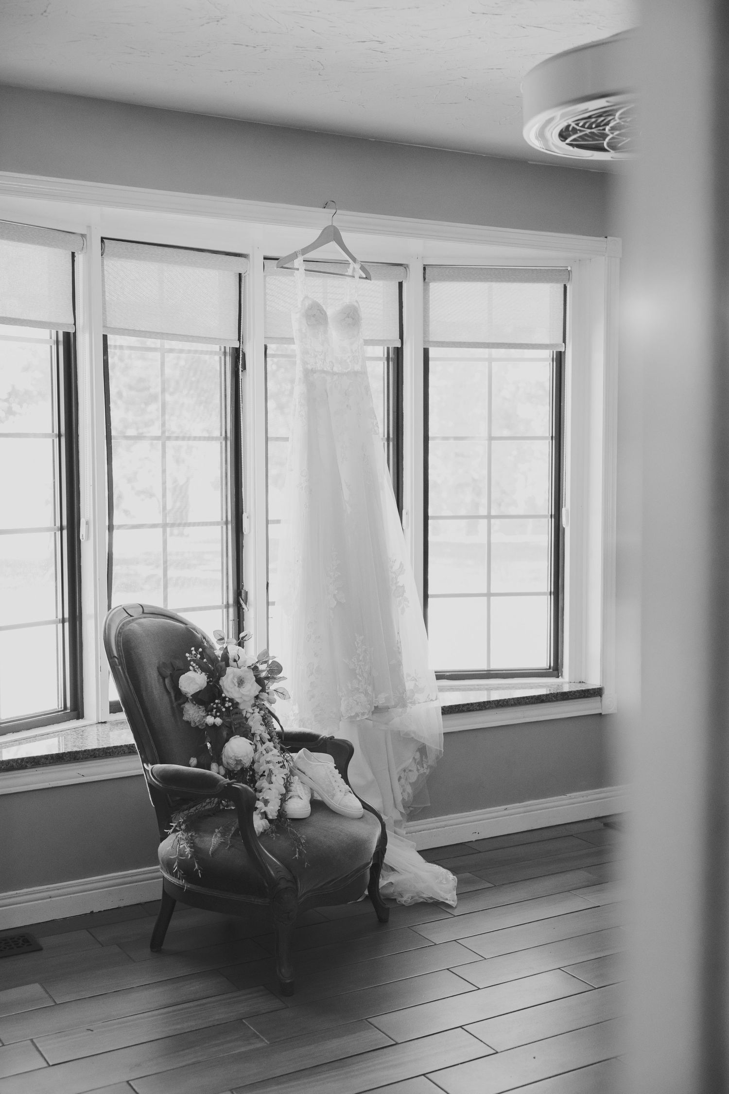 Black and white photo of a wedding dress hanging by a window above a vintage chair with a bridal bouquet.