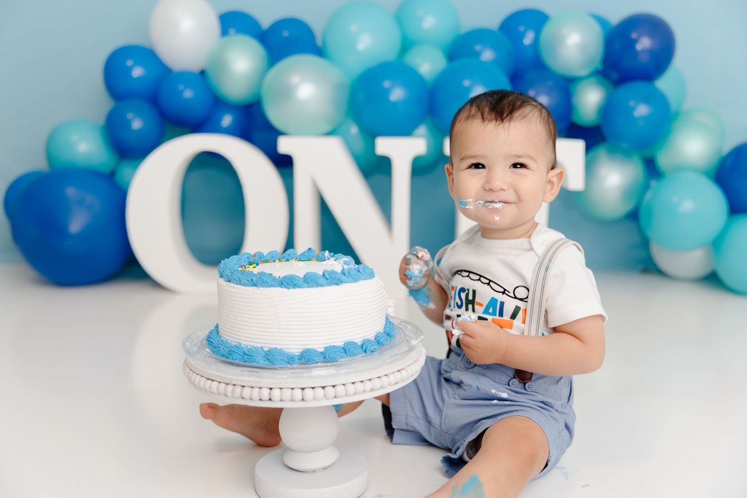 Smiling toddler celebrates first birthday with blue balloon arch and white cake decoration.
