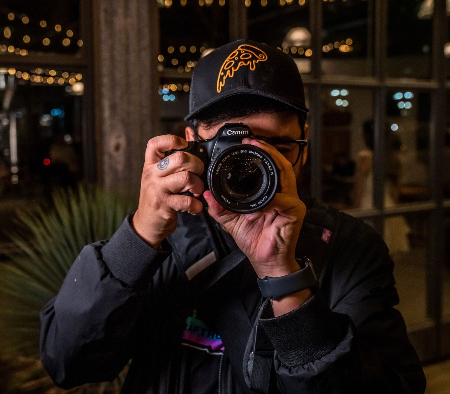 Photographer holding a Canon camera up to their face, wearing a black cap, at a night event with bokeh lights.