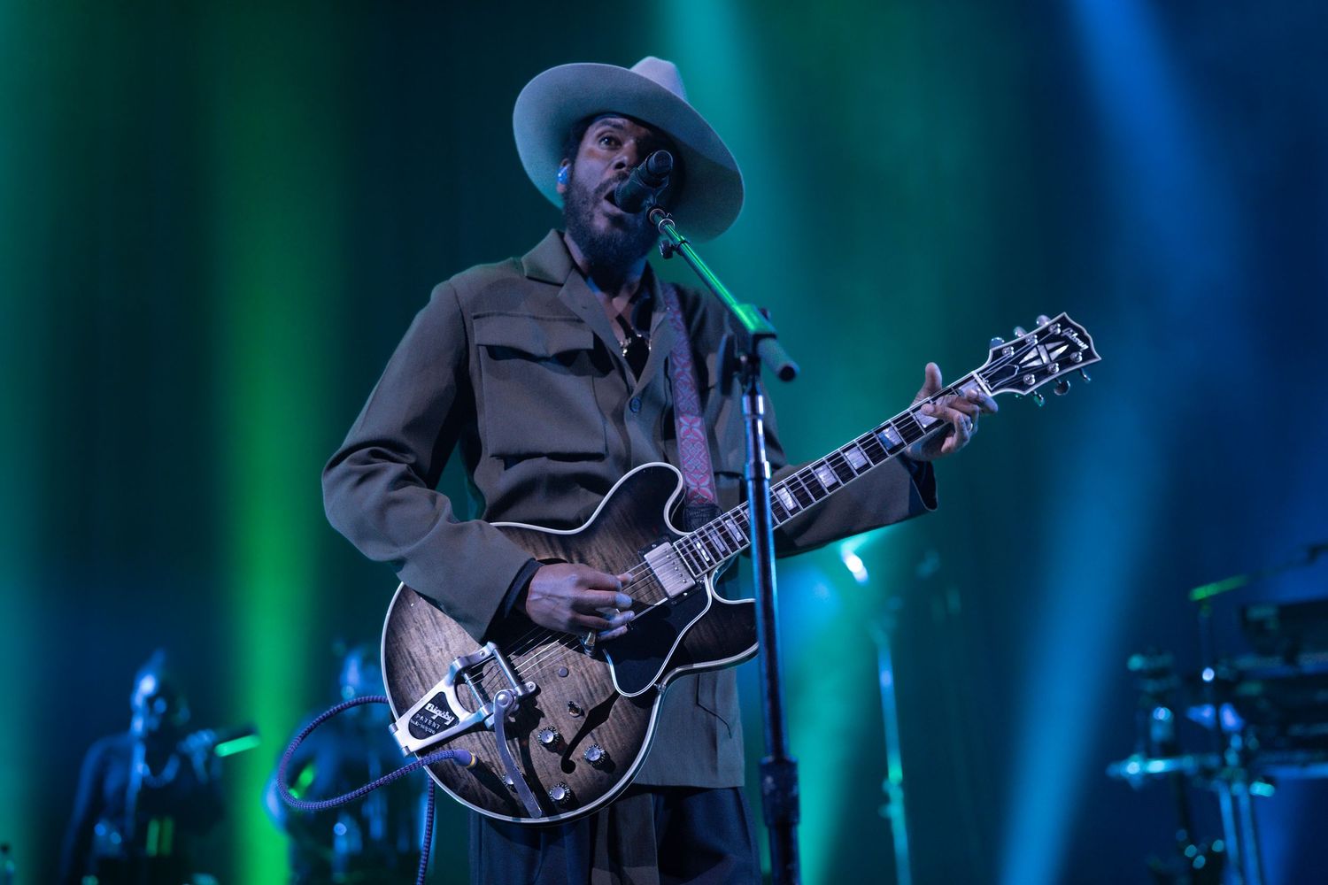Musician in cowboy hat playing hollow-body guitar on stage with green and blue lights.