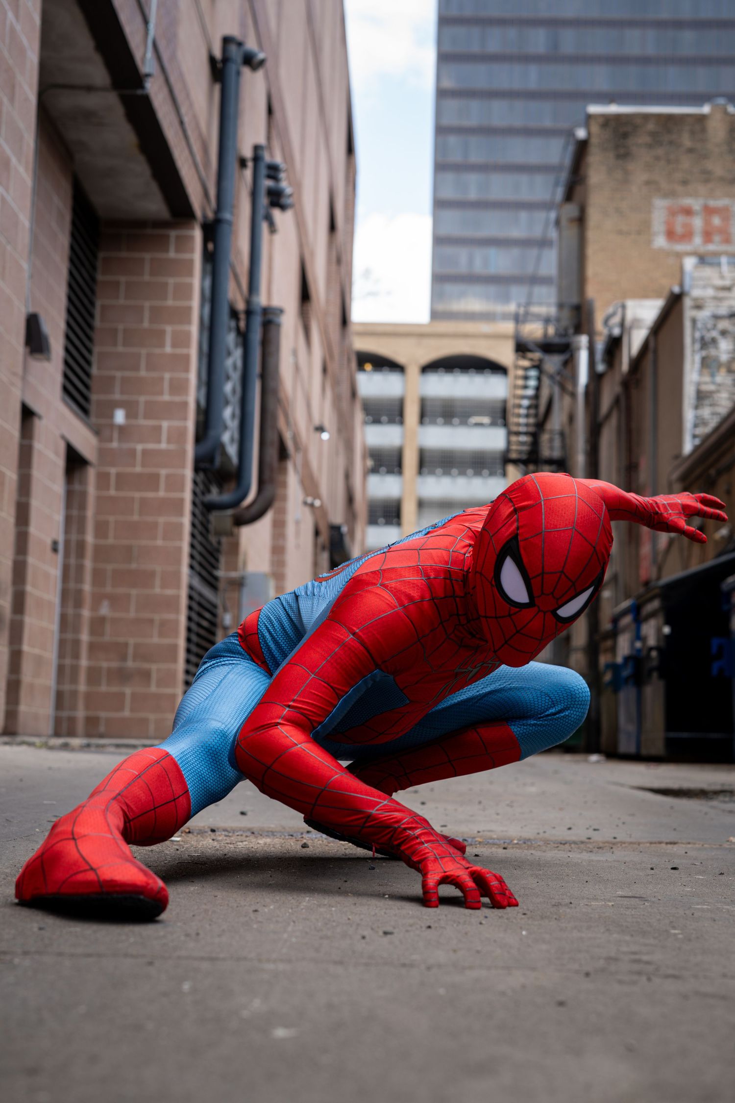 Person in a red and blue Spider-Man costume crouching in a urban alley between brick buildings.