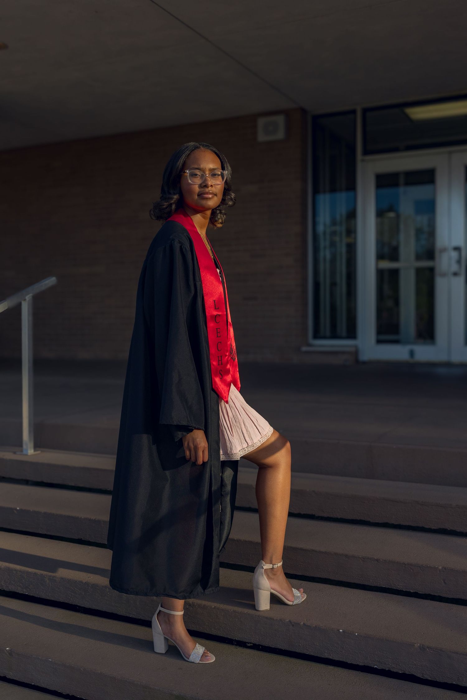 Young woman in black graduation gown with red stole and glasses poses on steps outside school building.