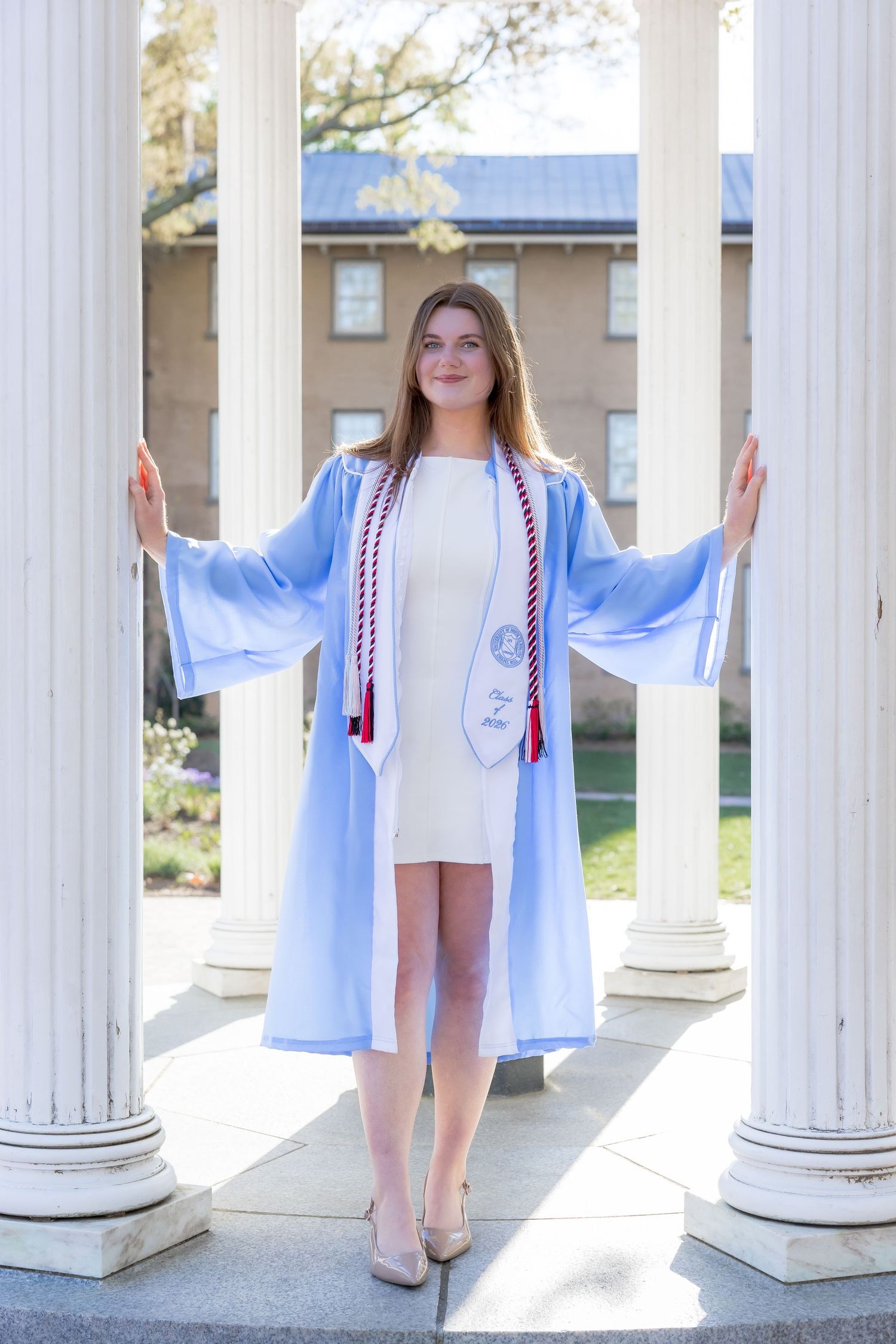 Young woman in light blue graduation gown and red stole poses between white columns at university campus.