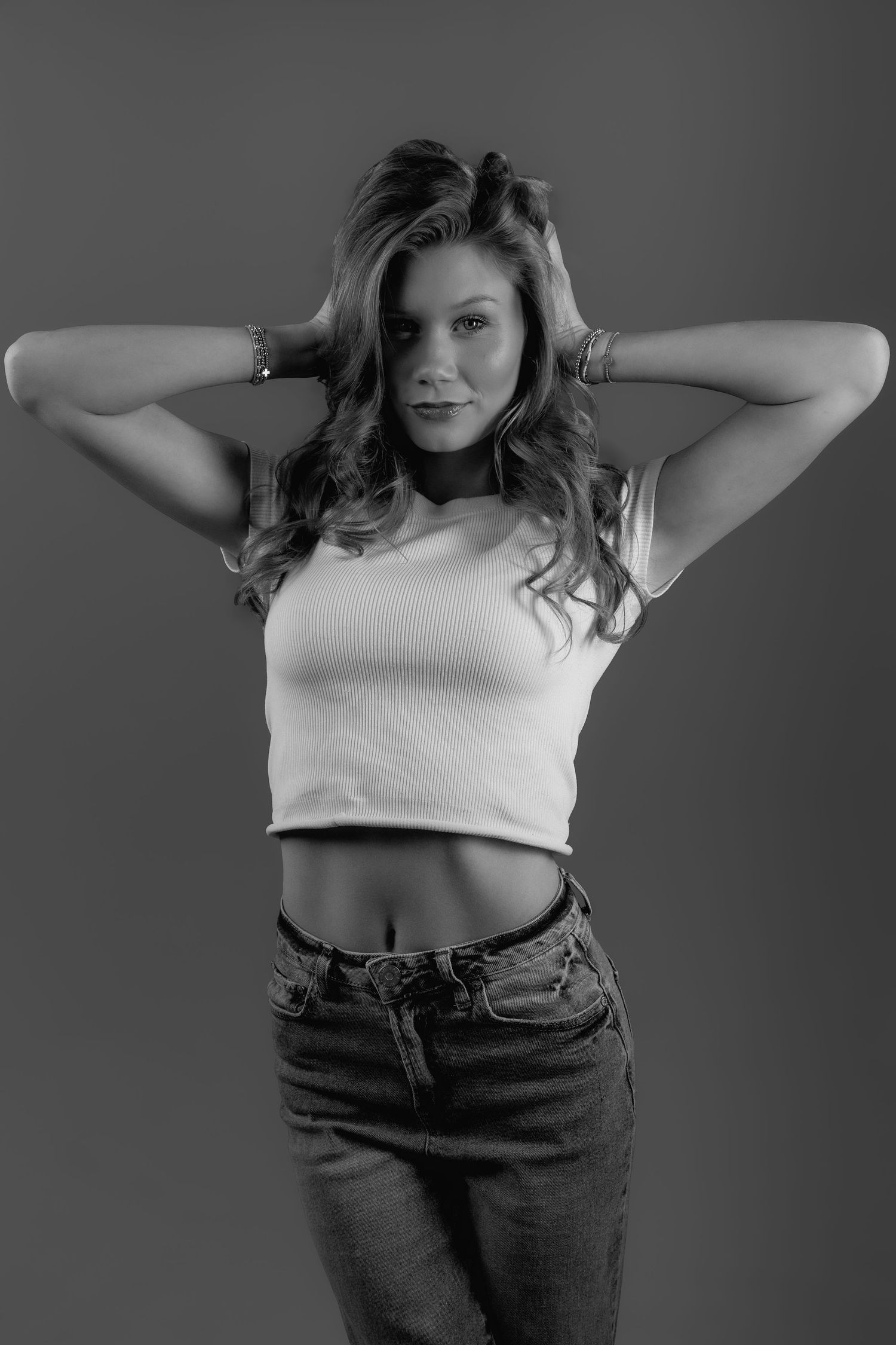 Black and white portrait of a young woman in crop top and jeans, hands behind head, posing against gray background.