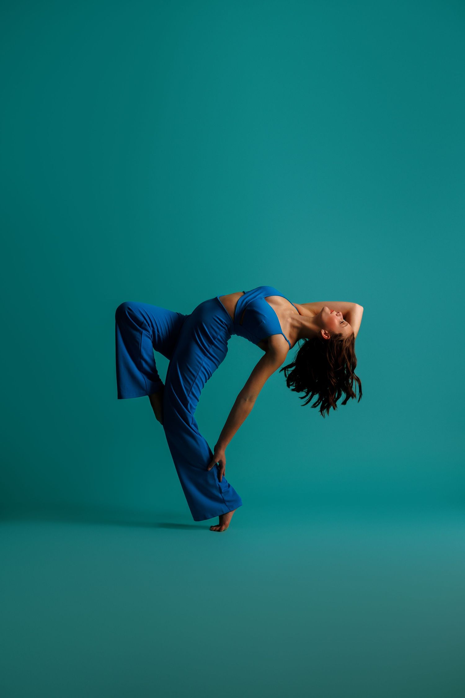 A dancer in blue wide-leg pants and crop top bends backward gracefully against a teal studio background.