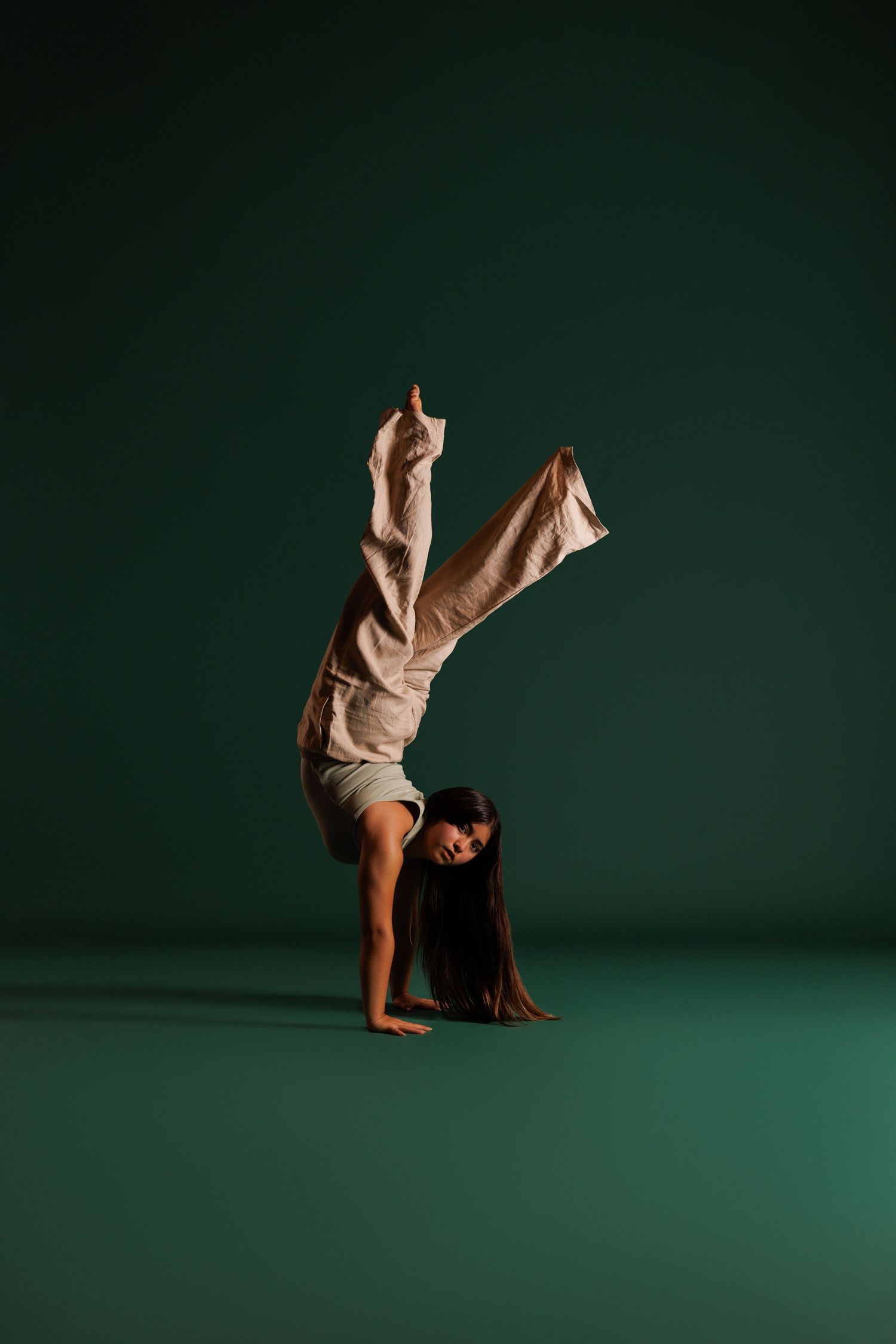 A dancer performs a handstand with legs split apart, wearing beige outfit against a dark green studio background.