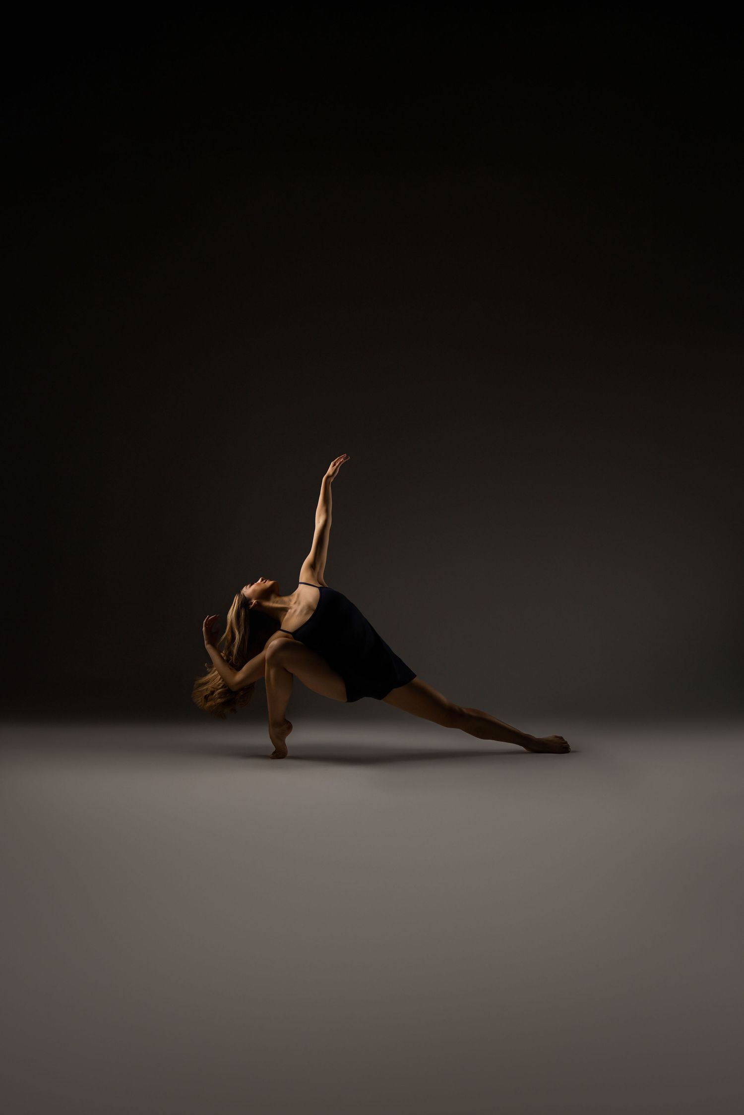 Contemporary dancer performs graceful movement sequence on dark stage floor with dramatic lighting and shadows.