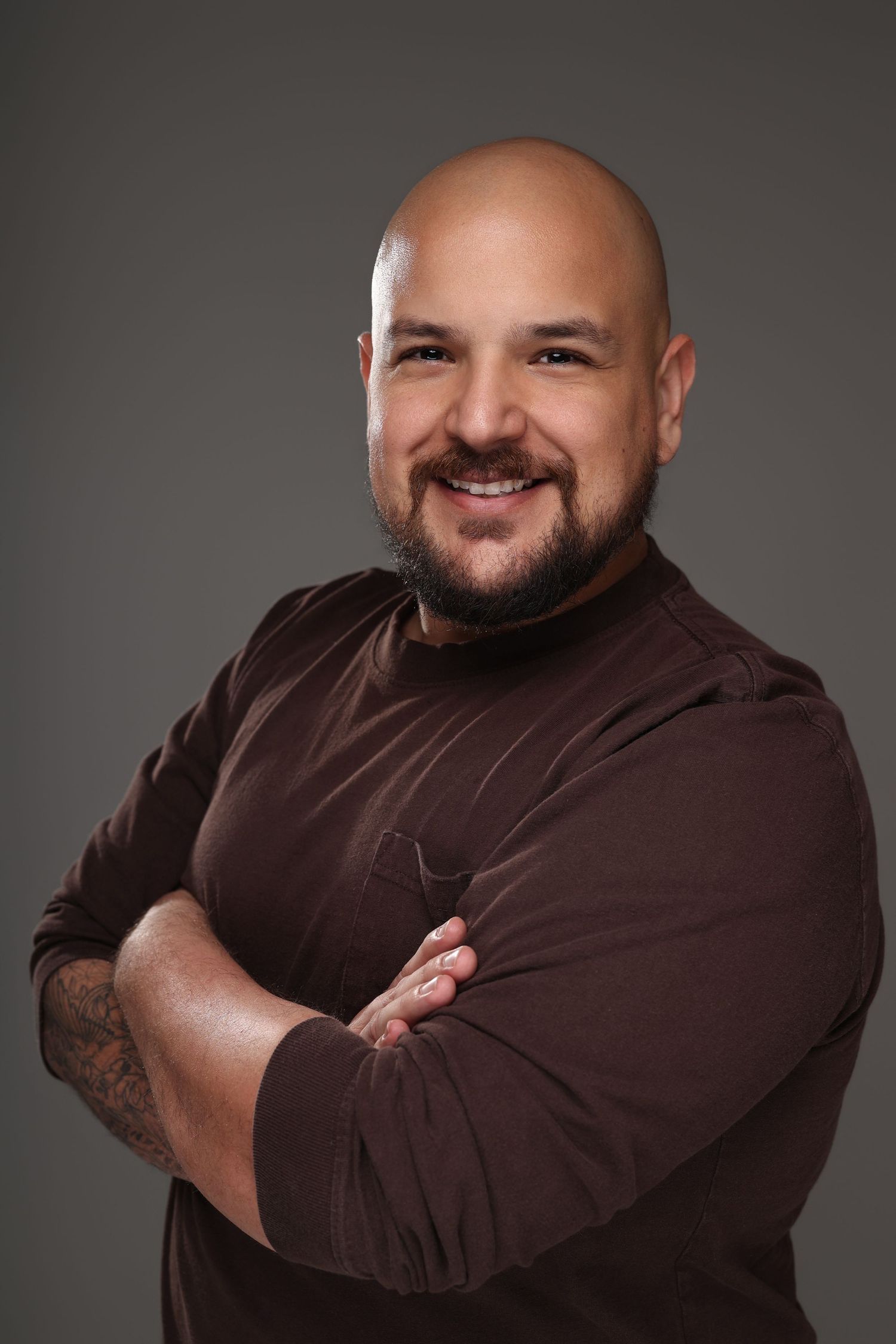 Smiling bald man with beard, arms crossed, wearing brown long-sleeve shirt, tattooed arm, gray background.
