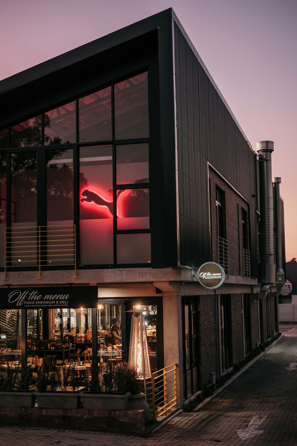 Modern black building at dusk housing Off The Menu restaurant with glowing Puma logo visible through large windows.