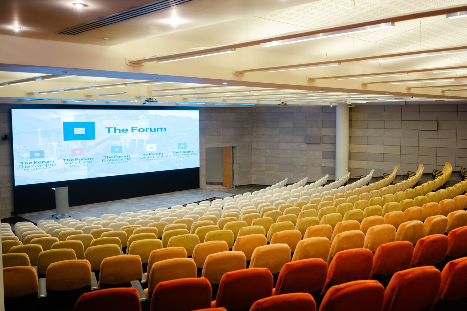 Empty conference hall with rows of orange seats facing a large screen displaying The Forum presentation.