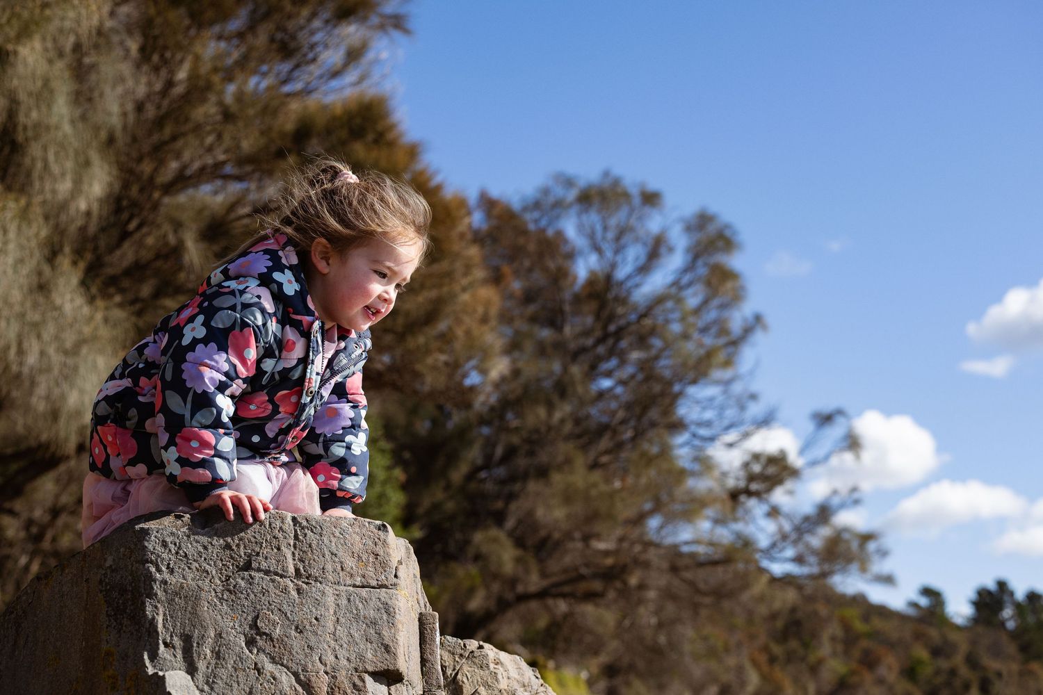 A child in a floral jacket leans over a large rock formation while exploring outdoors on a sunny day.