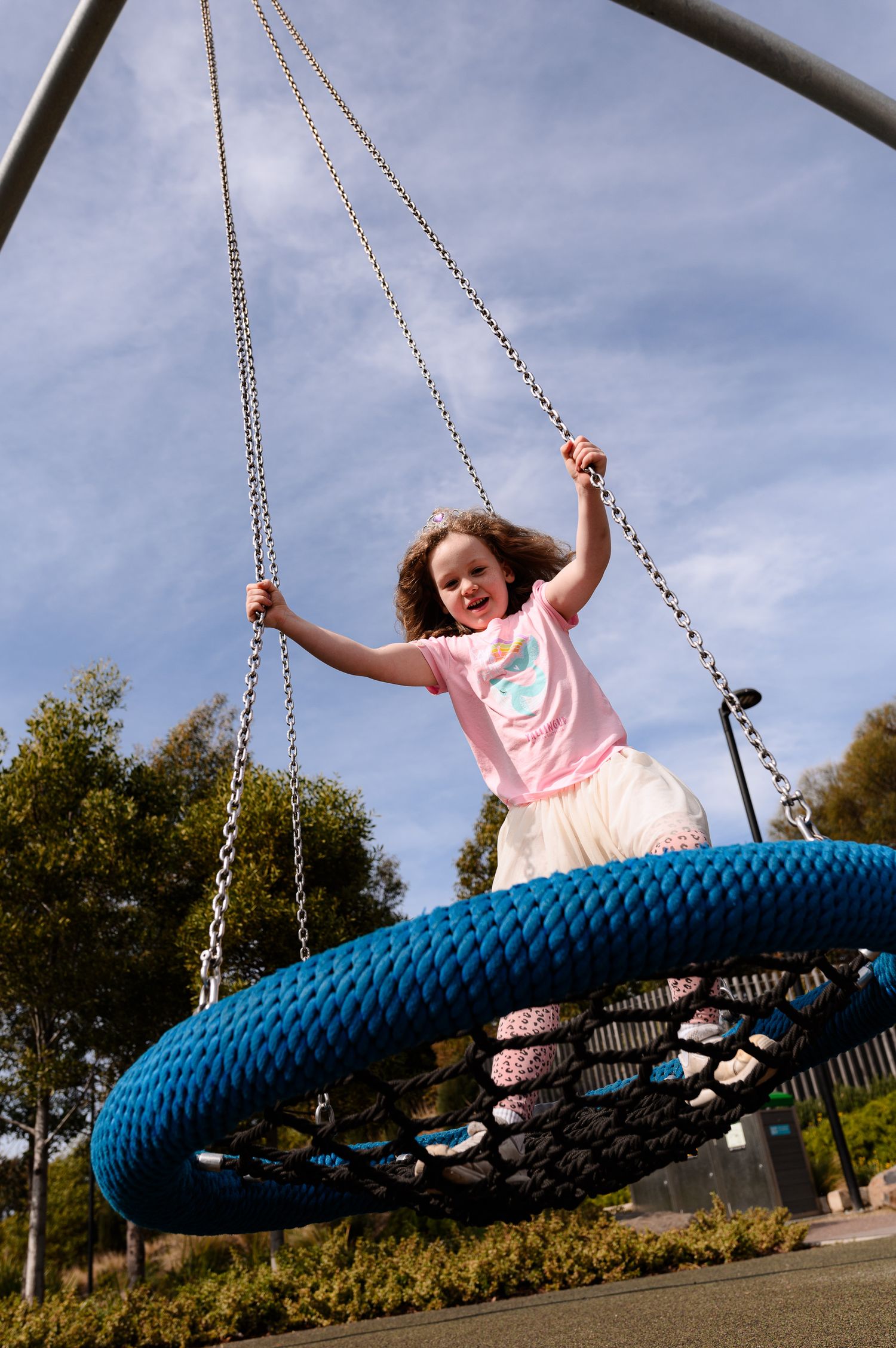 A person swings joyfully on a large blue whale-shaped playground swing against a bright blue sky.