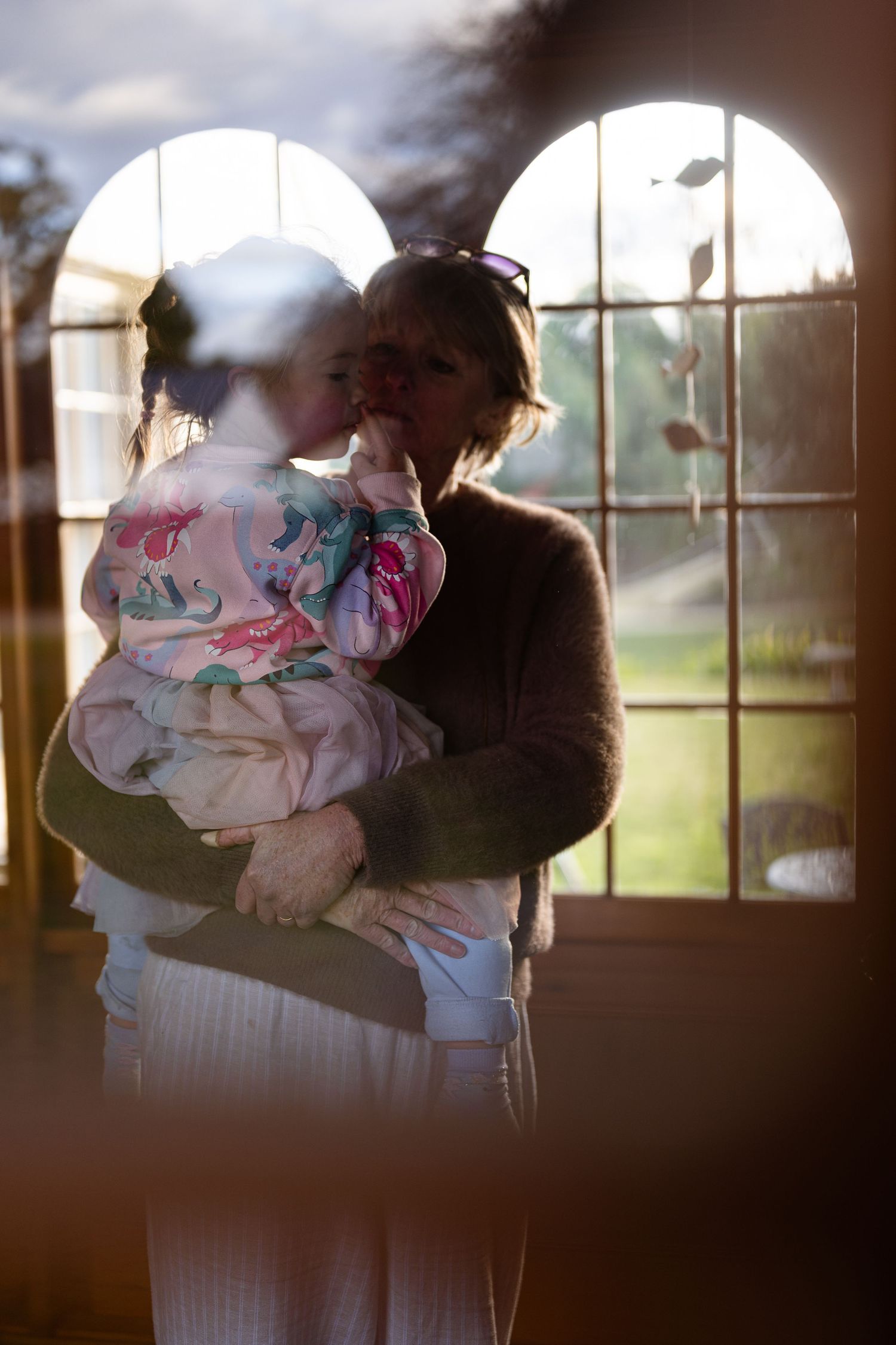 A silhouetted figure holds a small child wearing floral pajamas by a sunlit arched window.