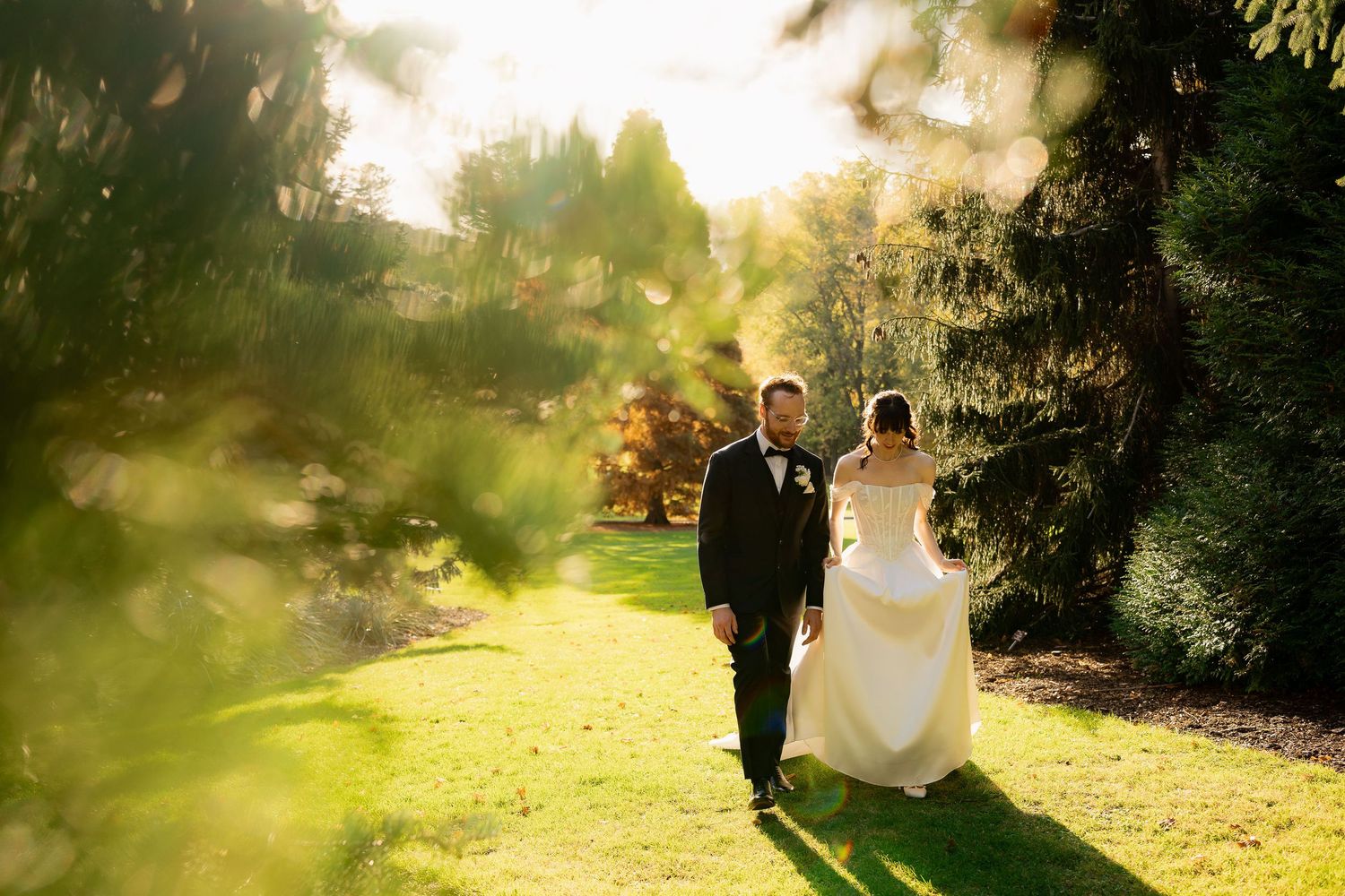 A couple walks through a sunlit garden path during golden hour with dreamy backlit lighting creating a romantic atmosphere.